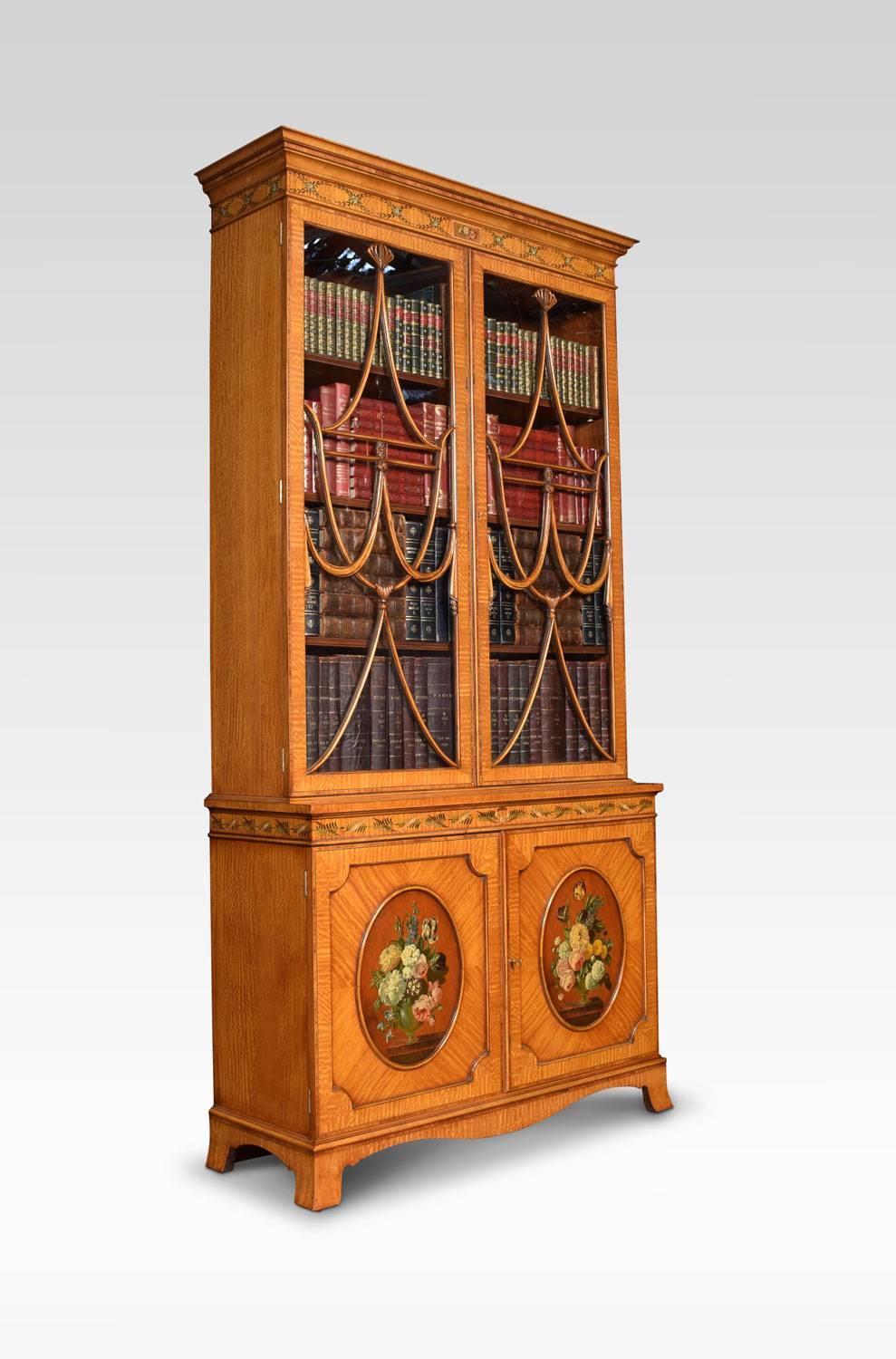 Satinwood bookcase with a flared cornice over foliated painted freeze. The two ornately beaded glazed doors enclosing adjustable shelved interior. The base section fitted with a pair of panelled cupboard doors having oval floral panels. All raised