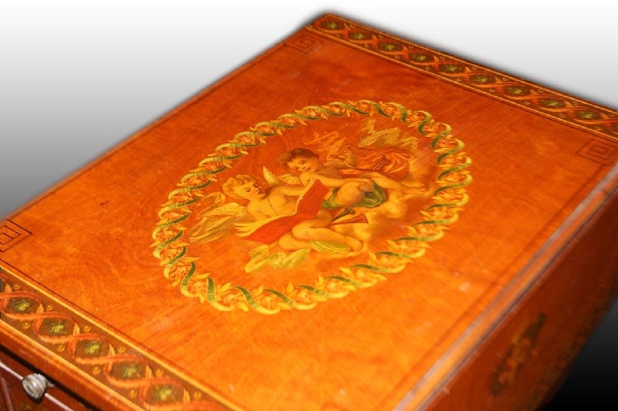 Beautiful Sheraton style English fliptop table from the first half of the 19th century, made of satinwood. It features a rich painting on the central top depicting two putti, with the edges of the top, legs, and drawers also richly painted with