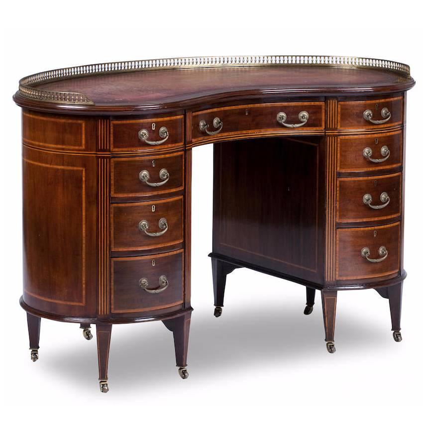 Sheraton style kidney shaped mahogany inlaid desk, circa 1900. The top has a three quarter wraparound brass gallery which has been gilded, and a faded burgundy leather writing surface which is gilded and tooled, surrounded by a crossbanding and