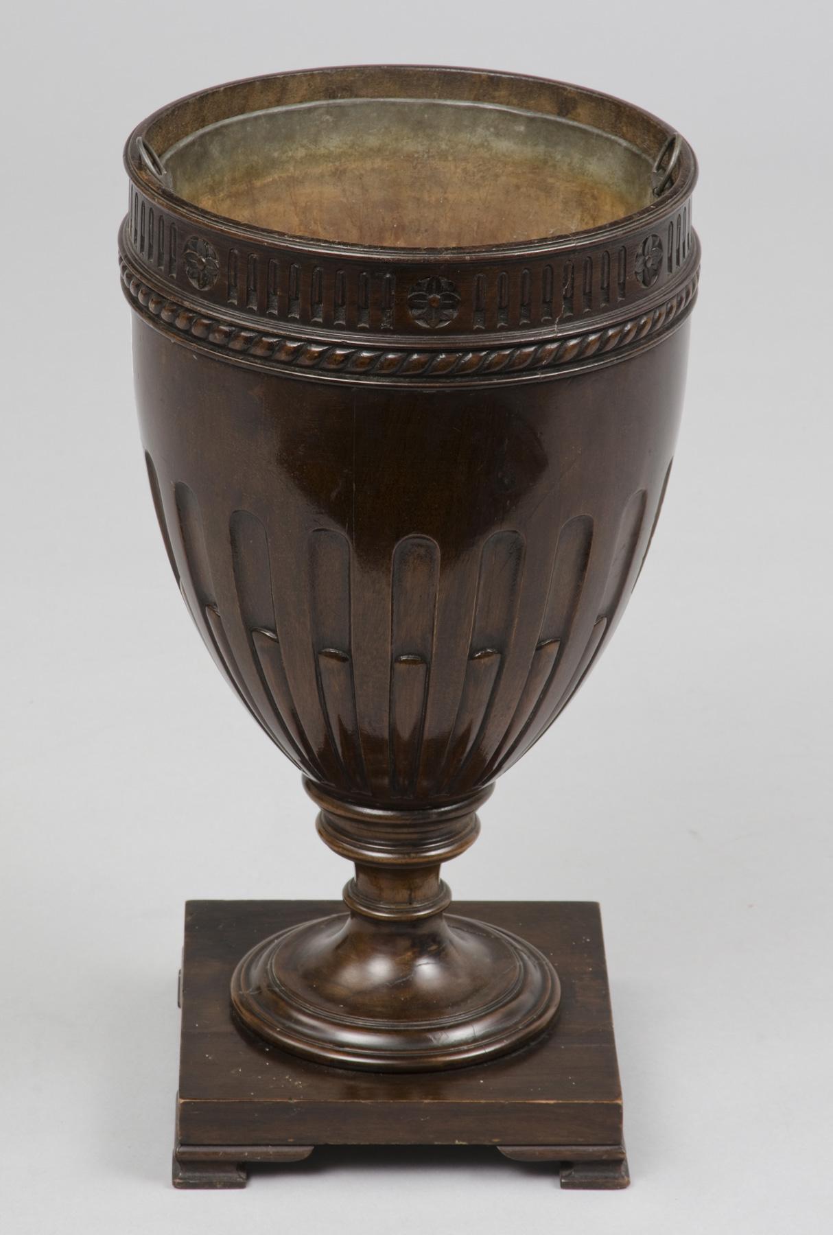 Sheraton style mahogany cutlery vase form urn, the top with carved fluting, stylized flowers and rope border, the bottom with carved stop fluting on turned pedestal and square base on bracket feet. Now converted to a jardinière with a metal liner.