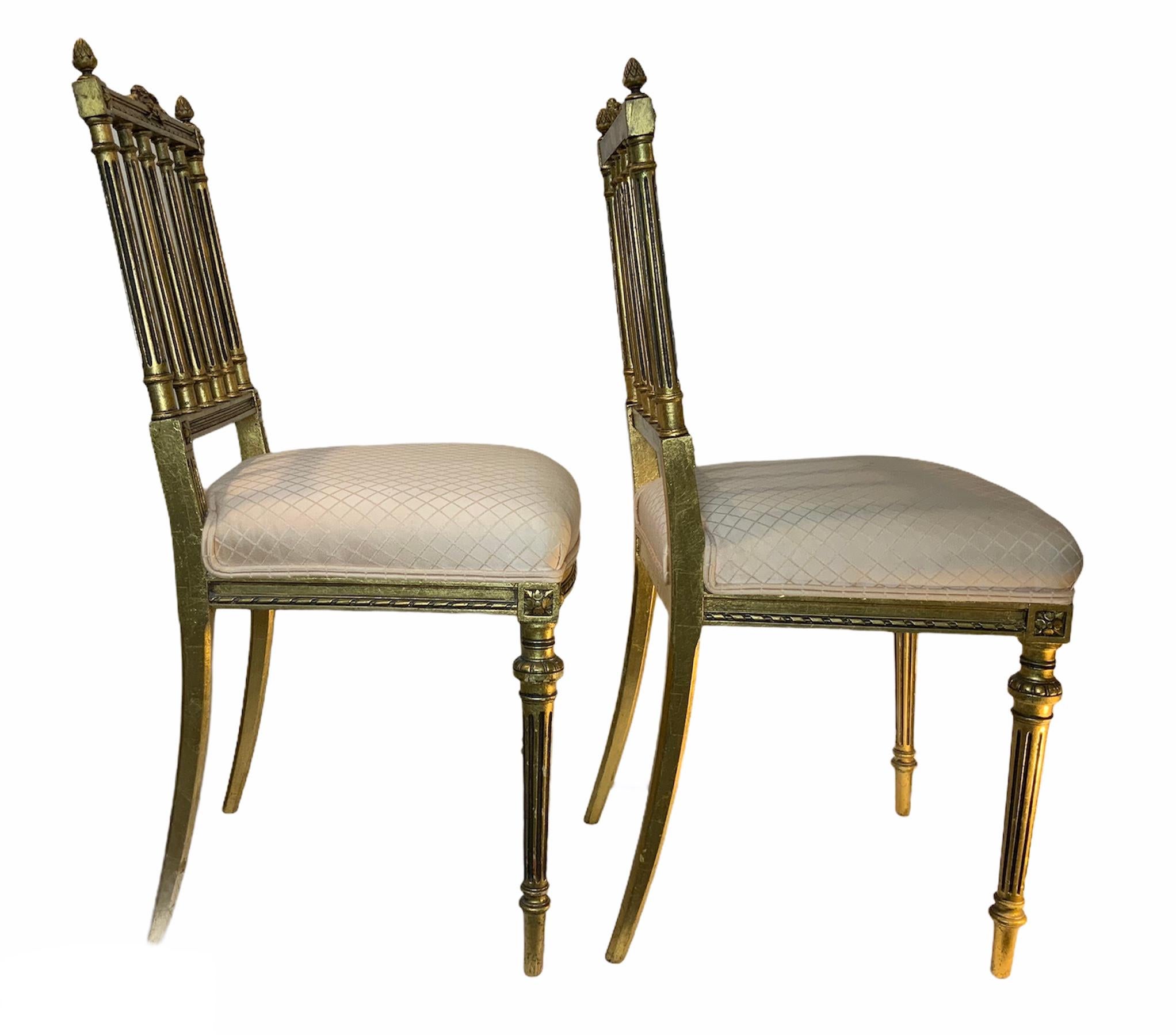 This is a pair of Sheraton style gilt wood chairs that have square back with six fluted balusters. The upper center borders of the chairs are adorned with a wreath of flowers that have a bow with two long legs of ribbons folded in each side of it.