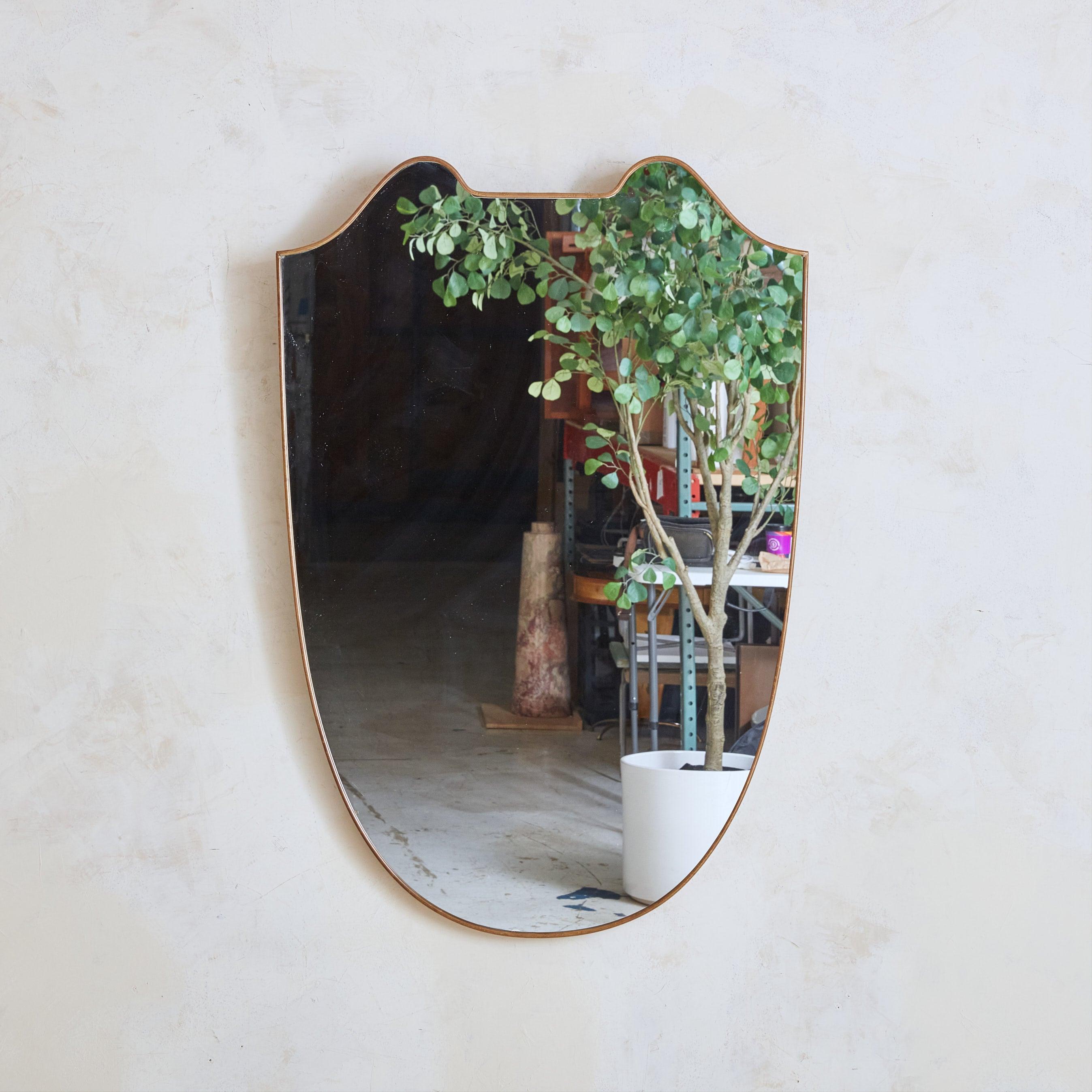 Specchio da parete italiano a forma di scudo degli anni '70, caratterizzato da eleganti curve lungo la parte superiore e da una silhouette sottilmente affusolata. Questo specchio ha una delicata cornice in ottone con una bellissima patina