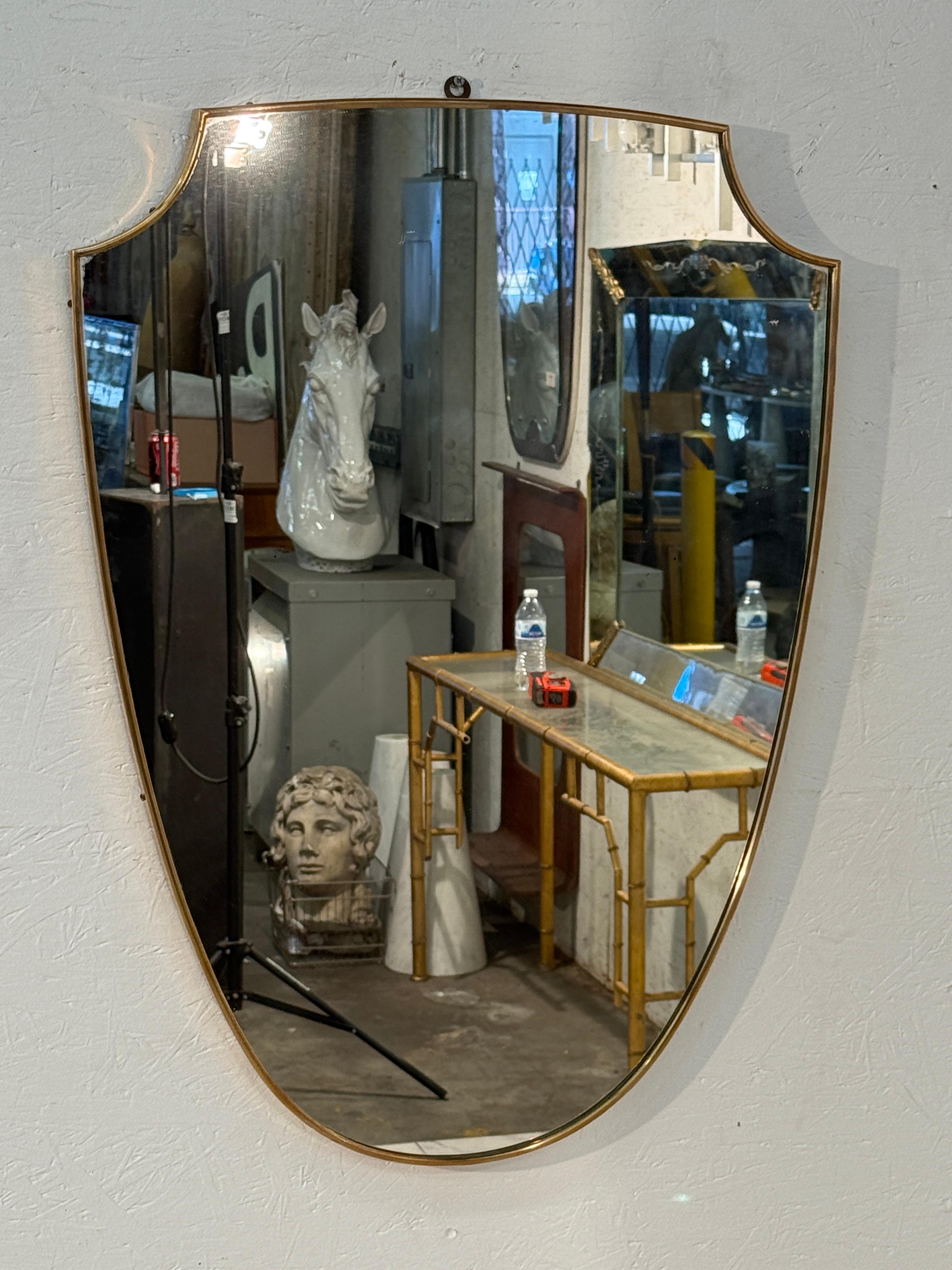 An Italian Mid Century  shield-shaped wall mirror framed in slim brass, combining classic form with refined simplicity. The unique silhouette adds character and a subtle sense of grandeur, while the polished brass edging introduces a warm metallic