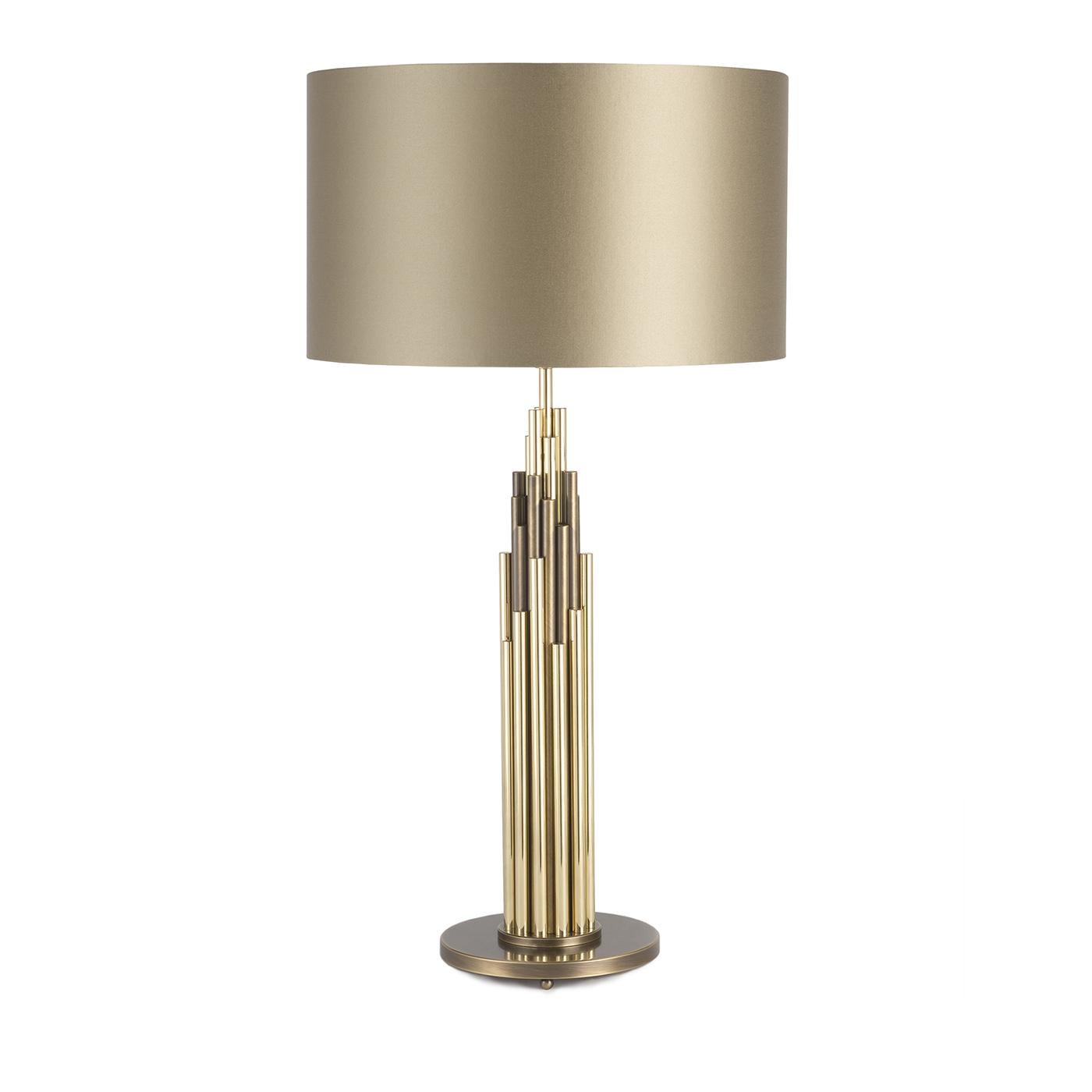 This elegant table lamp is part of the Shine collection and features a bronzed brass circular base supporting a series of vertical brass elements of different heights, some with a natural finish, some with a bronzed coloration. The satin shade above