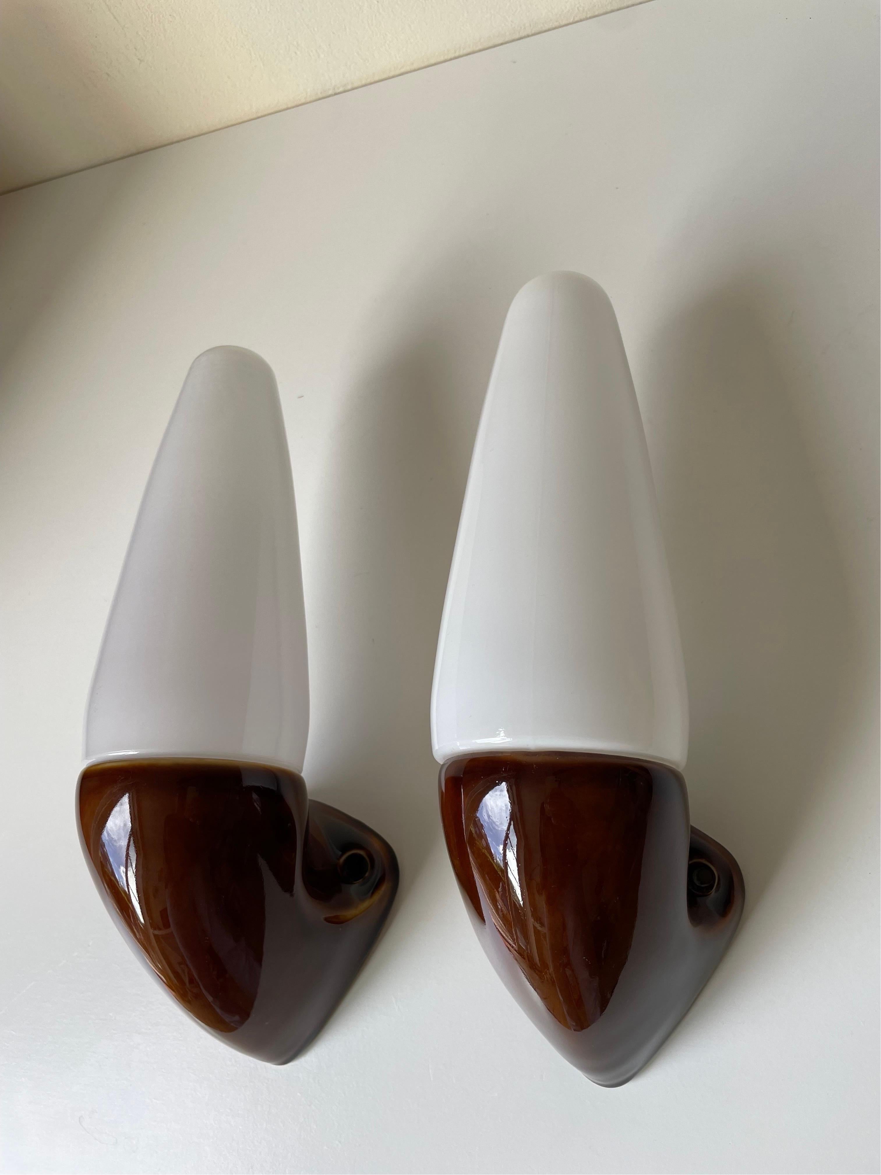 Set of two Swedish midcentury modern vintage wall sconces designed by artistic director for Ifö, Stig Carlsson (1932-2008) in the early 1960s. Shiny ochre brown glazed mount with cone shaped white opaline glass top. Original porcelain fitting for