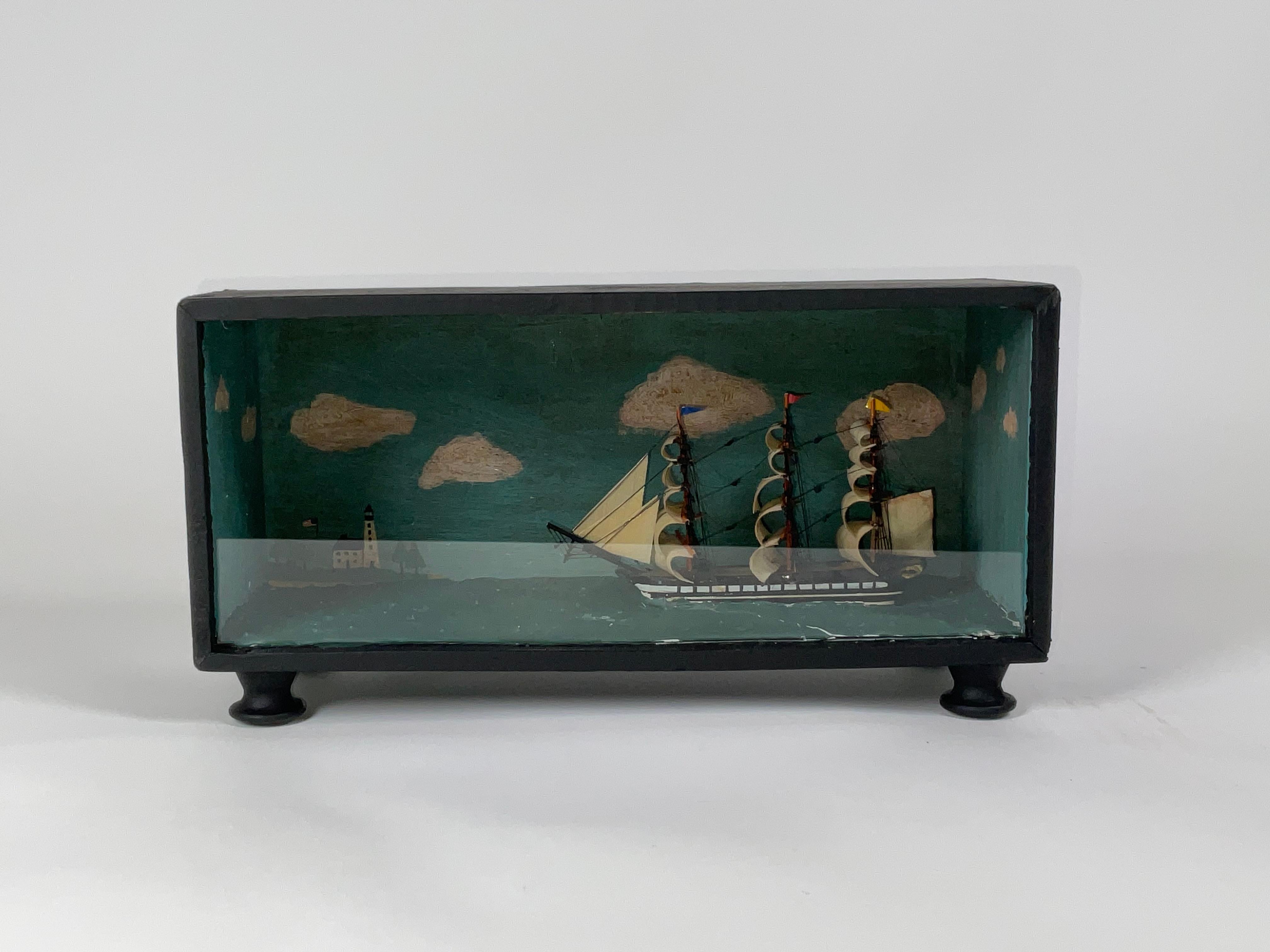 Diorama nautique d'un navire de guerre à gréement carré et à pleines voiles. Le bateau est posé sur sa ligne de flottaison, dans une mer calme, avec un arrière-plan peint d'un phare et de nuages. 6