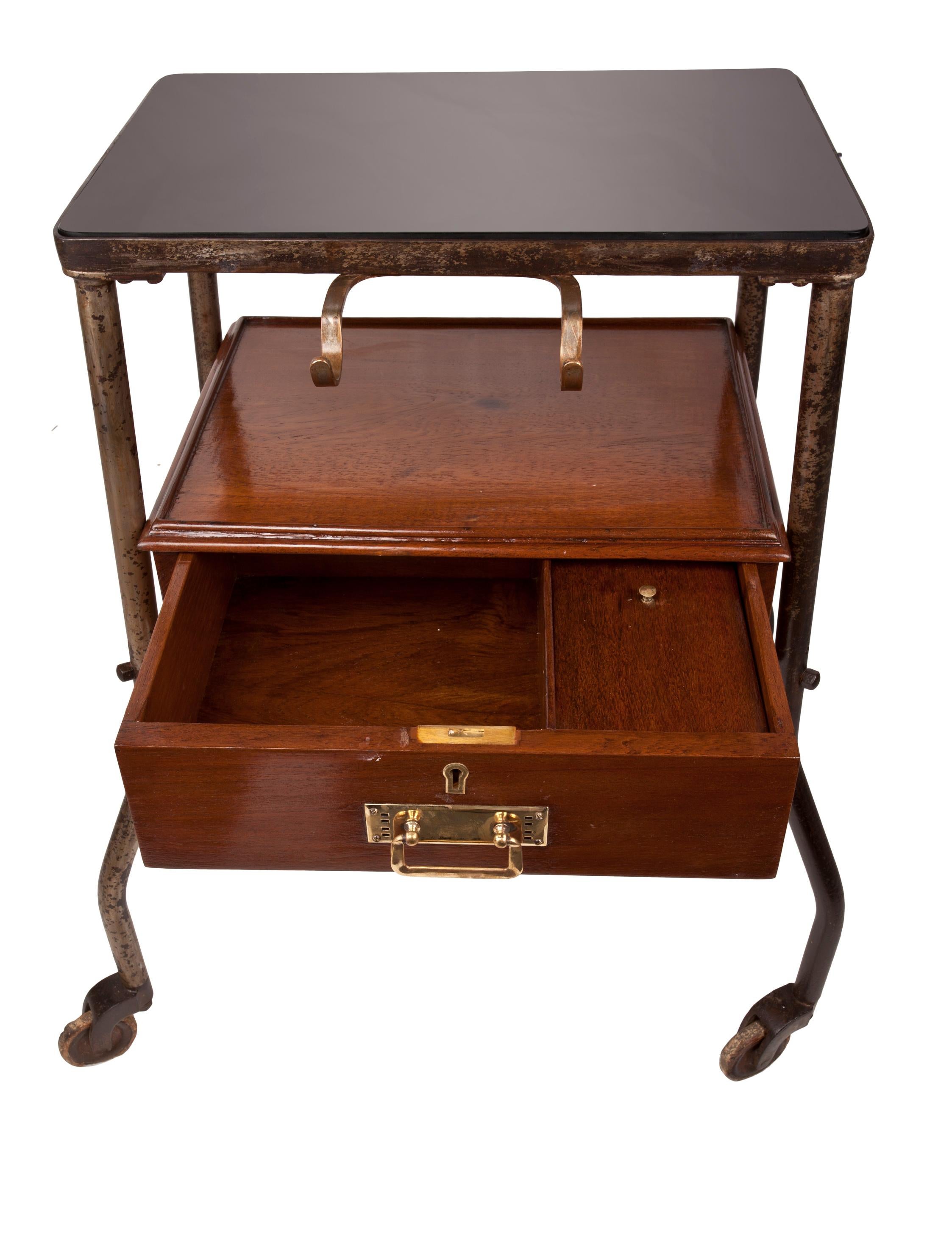 From a decommissioned ship, a teak medial trolley with smoked black glass top, front drawer and brass drawer pull and brass instrument holders in the front. Iron legs with the original iron wheels. 7 inches between the two levels. Great as a small