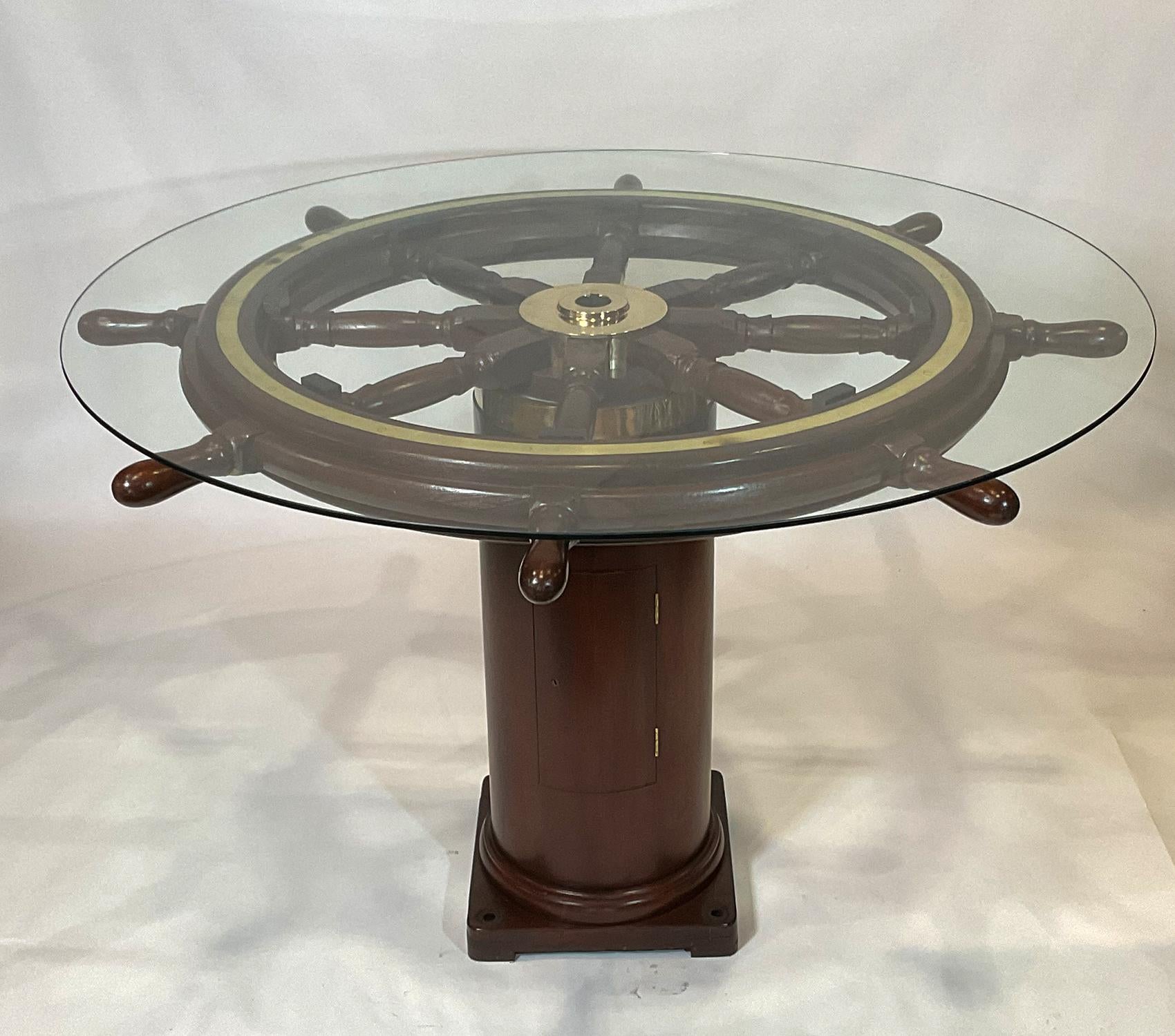 Fabulous antique ships wheel that has been fitted to a ships binnacle base creating a custom bar height dining table. This has a sixty-inch half inch tempered glass top. The eight-spoke wheel has been expertly refinished and is fitted with a highly