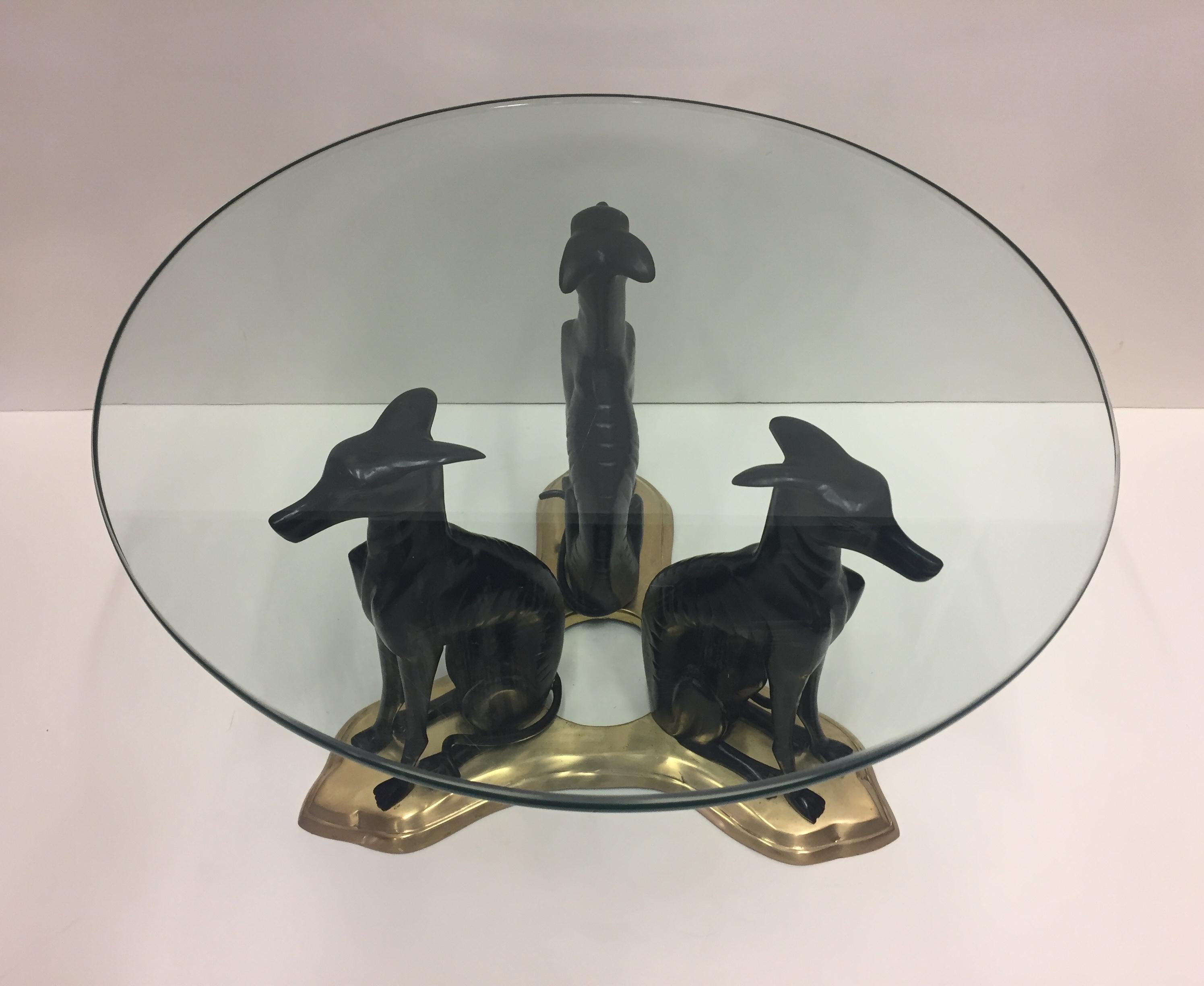 Dramatic round coffee table having an amazing base of 3 patinated bronze whippet dogs seated on bronze platform. Chunky thick glass top.