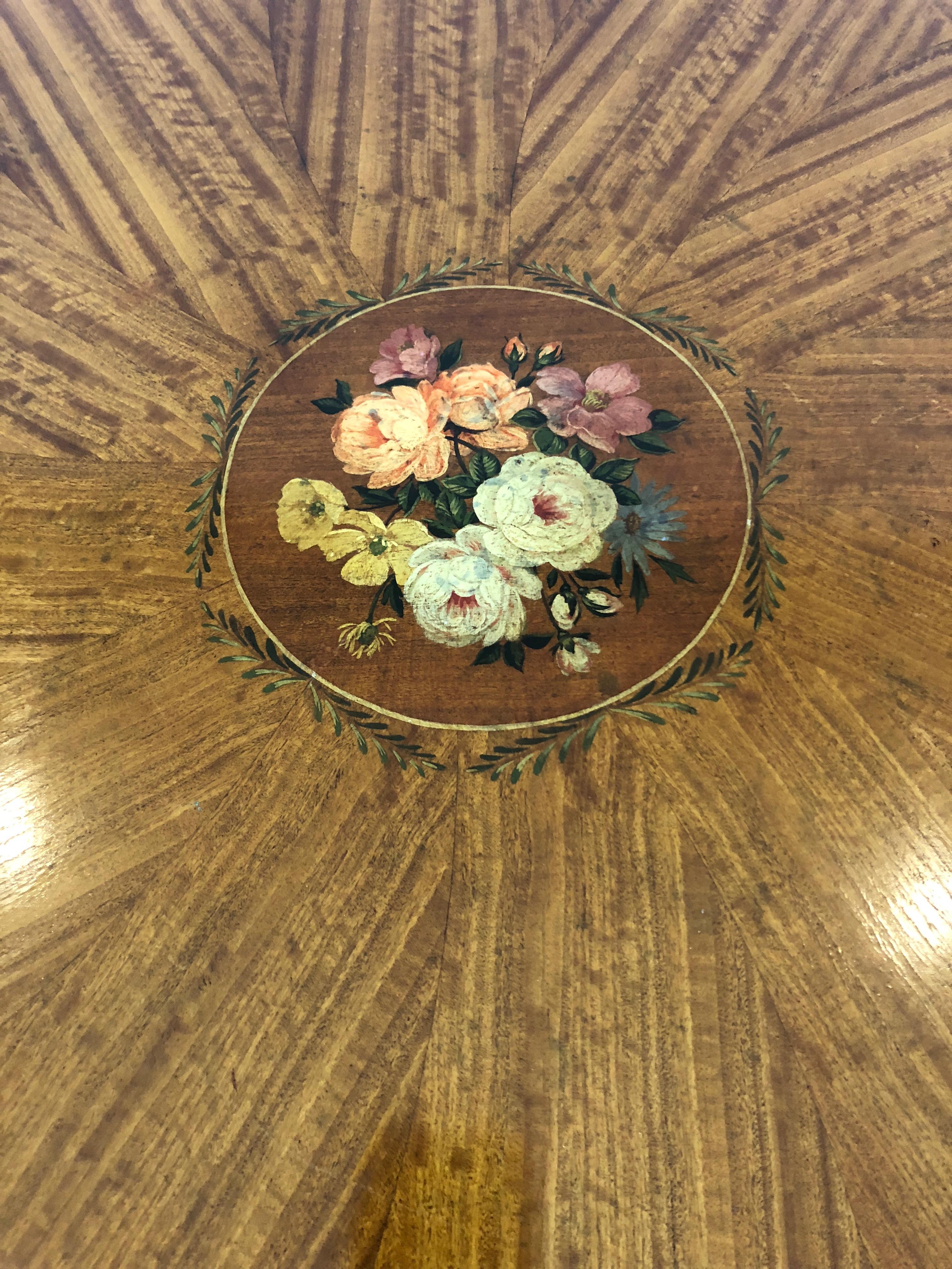 A gorgeous satinwood round centre table having a top where the wood is inlaid to look like the petals of a flower surrounding a central mandala of painted roses, resting on an elegant pedestal with four curved feet and brass caps and casters.
