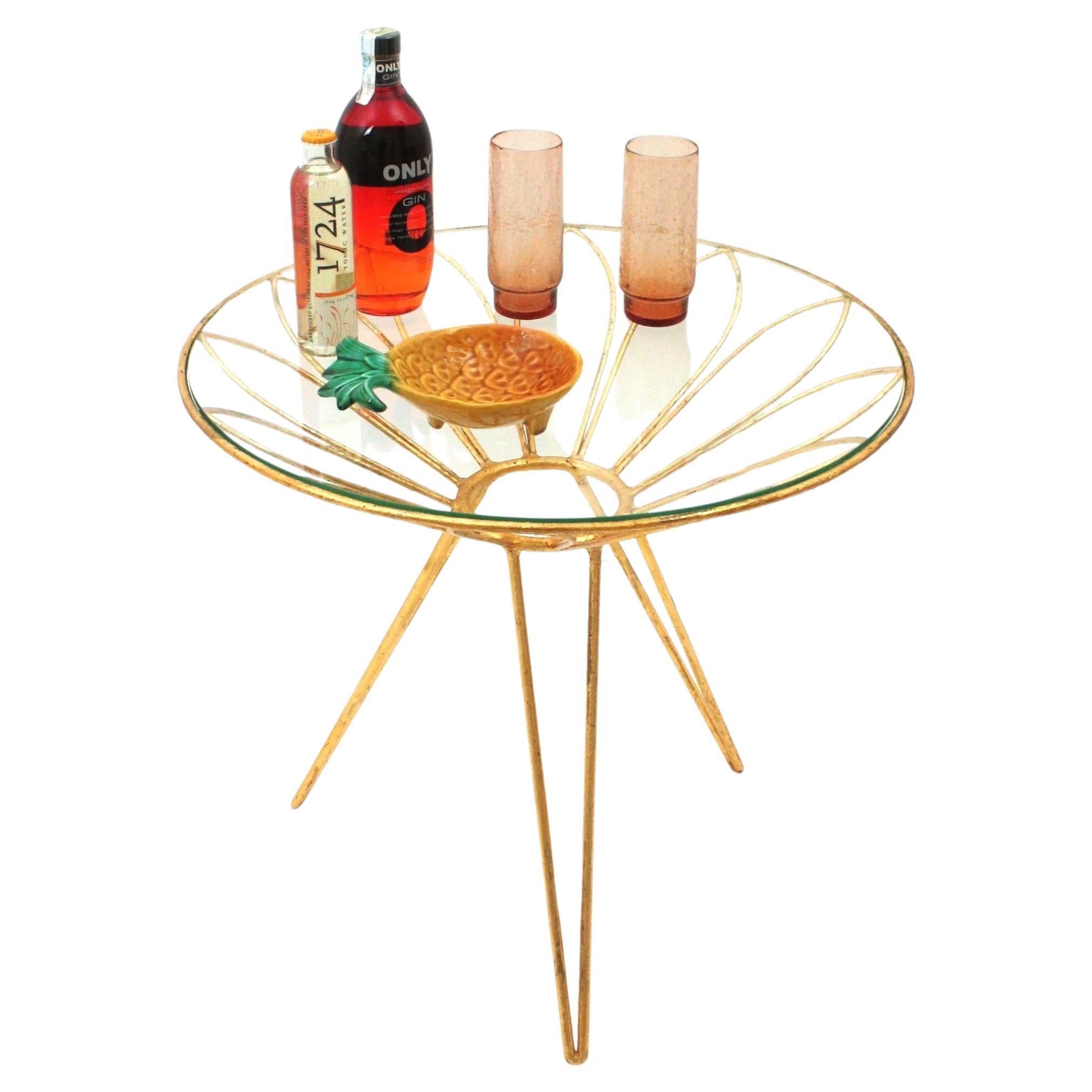 Tavolino da caffè Hollywood Regency a fiori di margherita in ferro dorato, Francia, 1950.
Tavolino glamour in ferro dorato con piano rotondo in vetro su base a treppiede. 
Questo tavolino da cocktail presenta un piano a forma di fiore di margherita