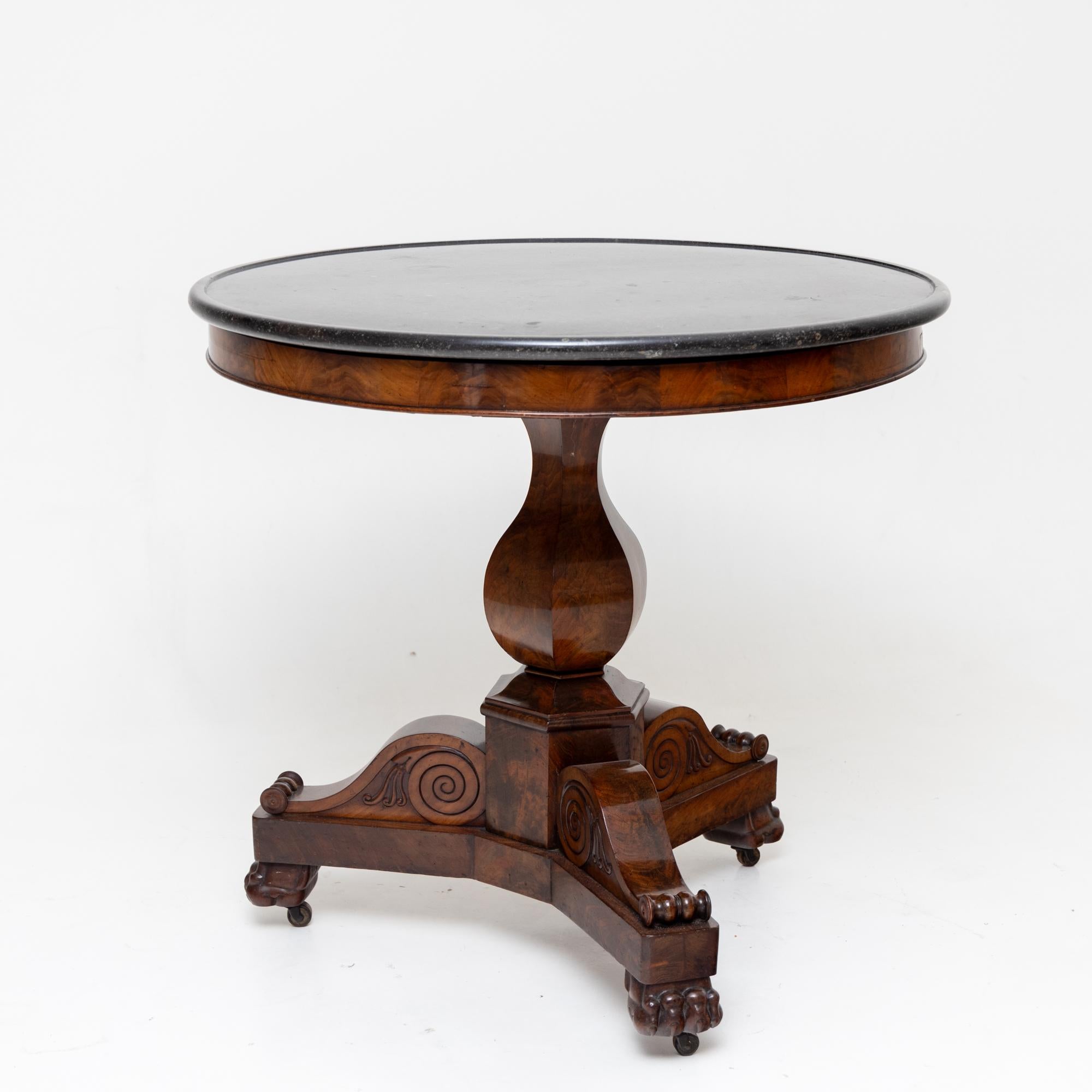French side table on a three-legged base with lion paws and elaborately carved volutes. Above it rises a hexagonal, tulip-shaped shaft. The round granite top in dark grey rests on an elegant smooth frame with a discreet moulding. Small brass castors