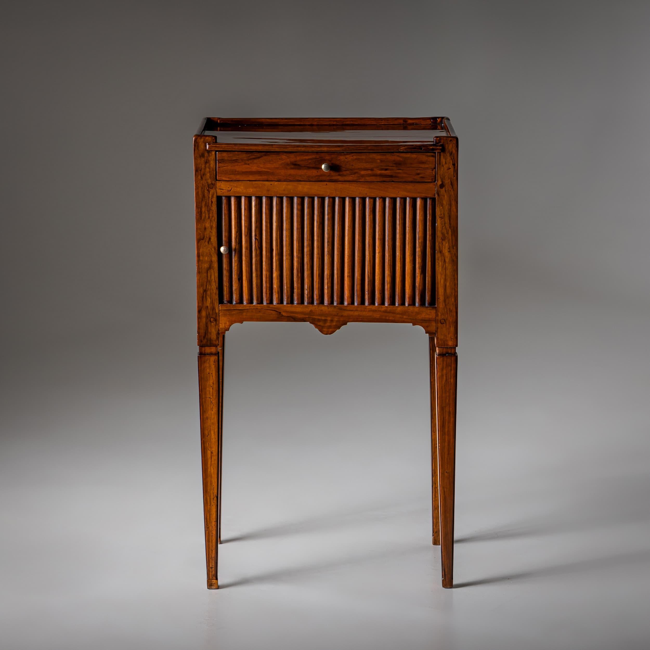 Small side table standing on tall, tapered square legs with an accentuated apron and a narrow drawer. The door is designed as a tambour door and slides open to the right. Crafted from fine walnut, this piece can also be used as a bedside table.