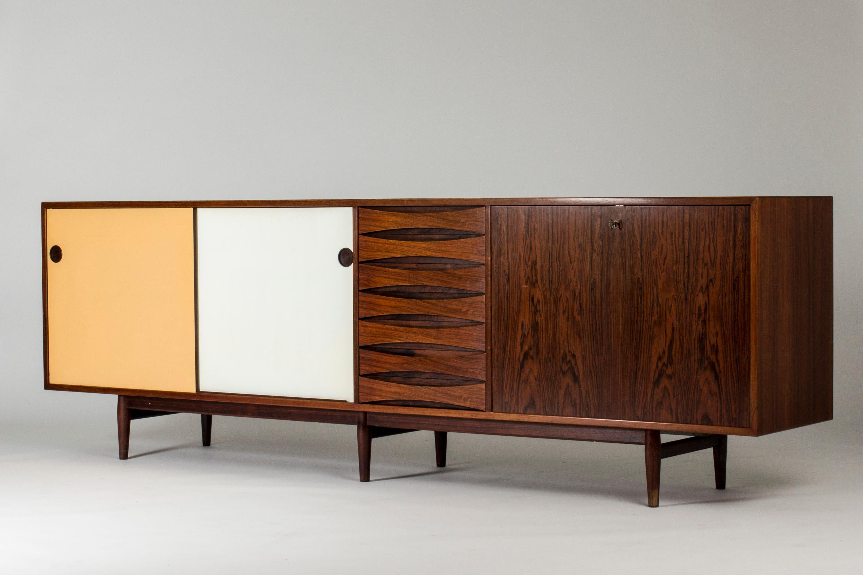 Striking rosewood sideboard by Arne Vodder, where yellow and white panel doors contrast with the cleanly sculpted drawers in the middle and the flaming rosewood veneer on the flap door on the left. The back of the sideboard is made with rosewood