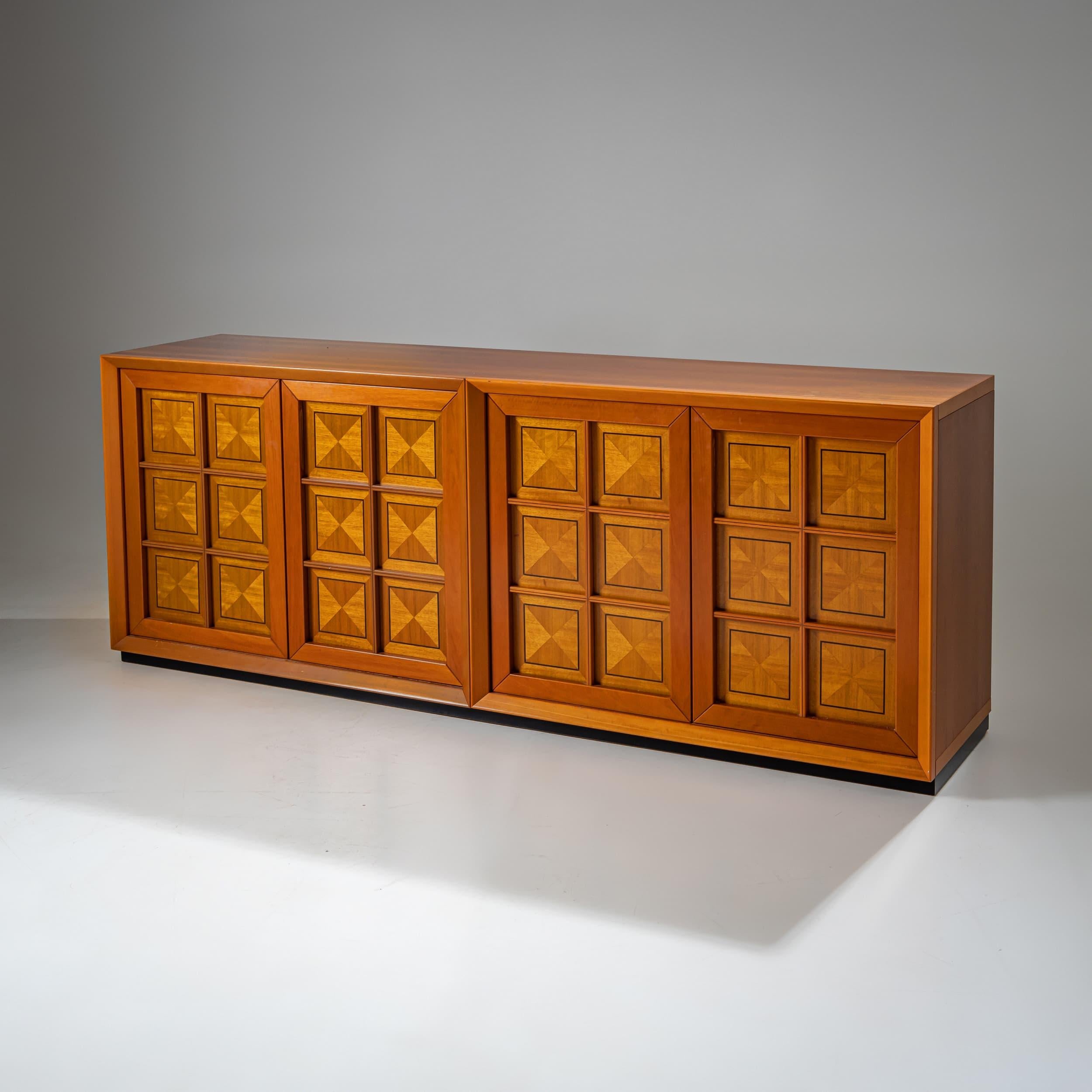 Aparador rectangular con un diseño limpio y lineal y llamativos detalles decorativos. Las cuatro puertas presentan incrustaciones de motivos cuadrados en madera clara y oscura, creando un efecto tridimensional. Los marcos biselados de las puertas se