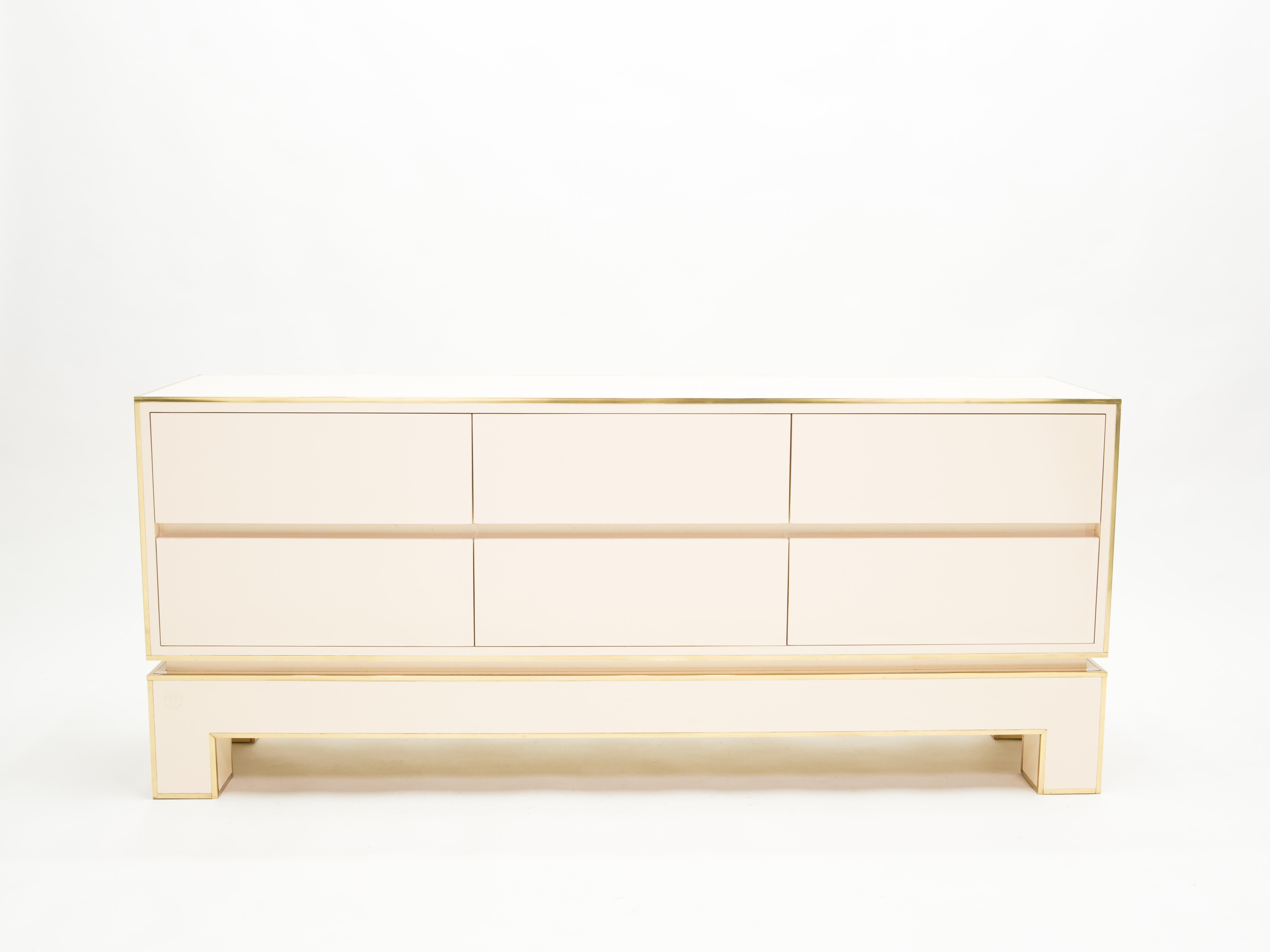 Crispy white lacquer, paired with bright brass accents, feels crisp and luxe on this beautiful and large commode / chest of drawers / sideboard. This beautiful and rare piece is signed by Alain Delon and edited by Maison Jansen in 1975. Its boxy,