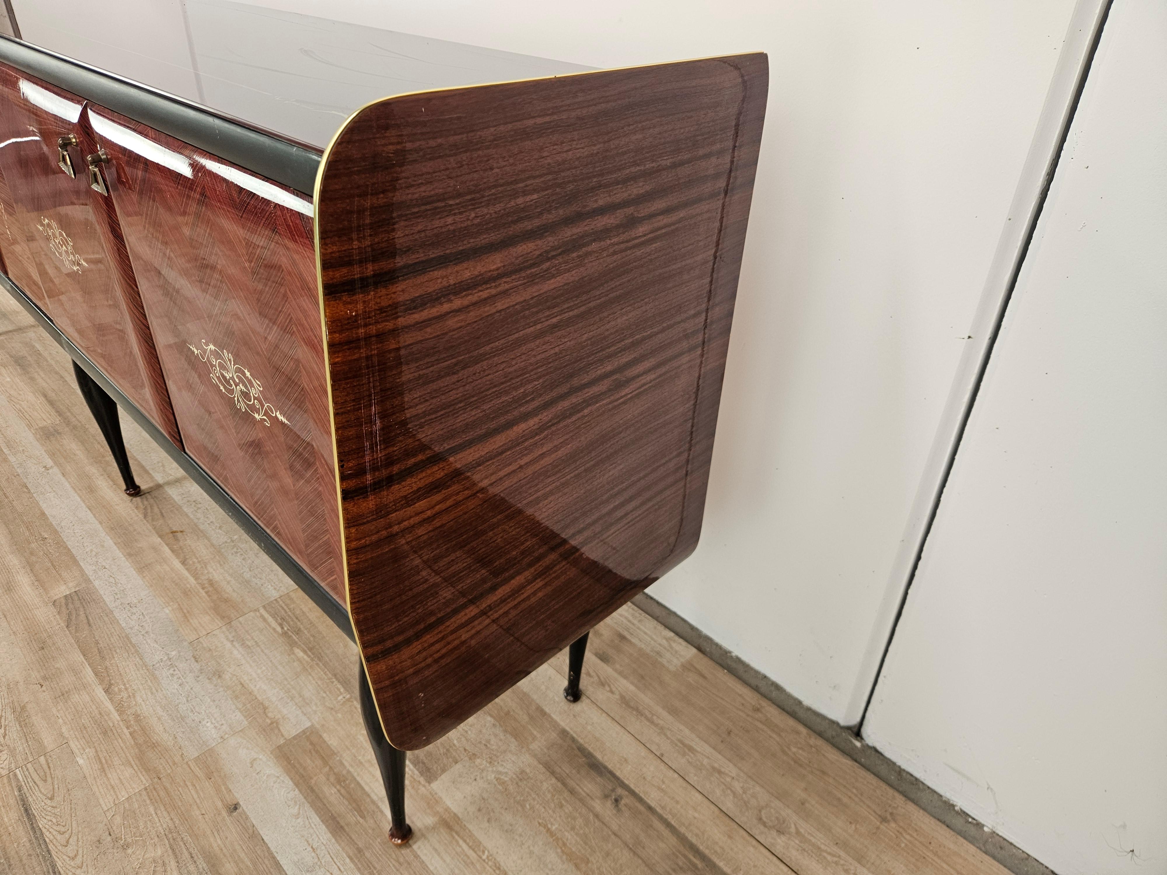 Elegante credenza vintage anni '50 rivestita in mogano con profili in ottone decorato e lavorato, molto fine e con un design moderno e vivace.

La credenza si presenta con un bellissimo vetro decorato sul piano, dispone di ante laterali con ripiano