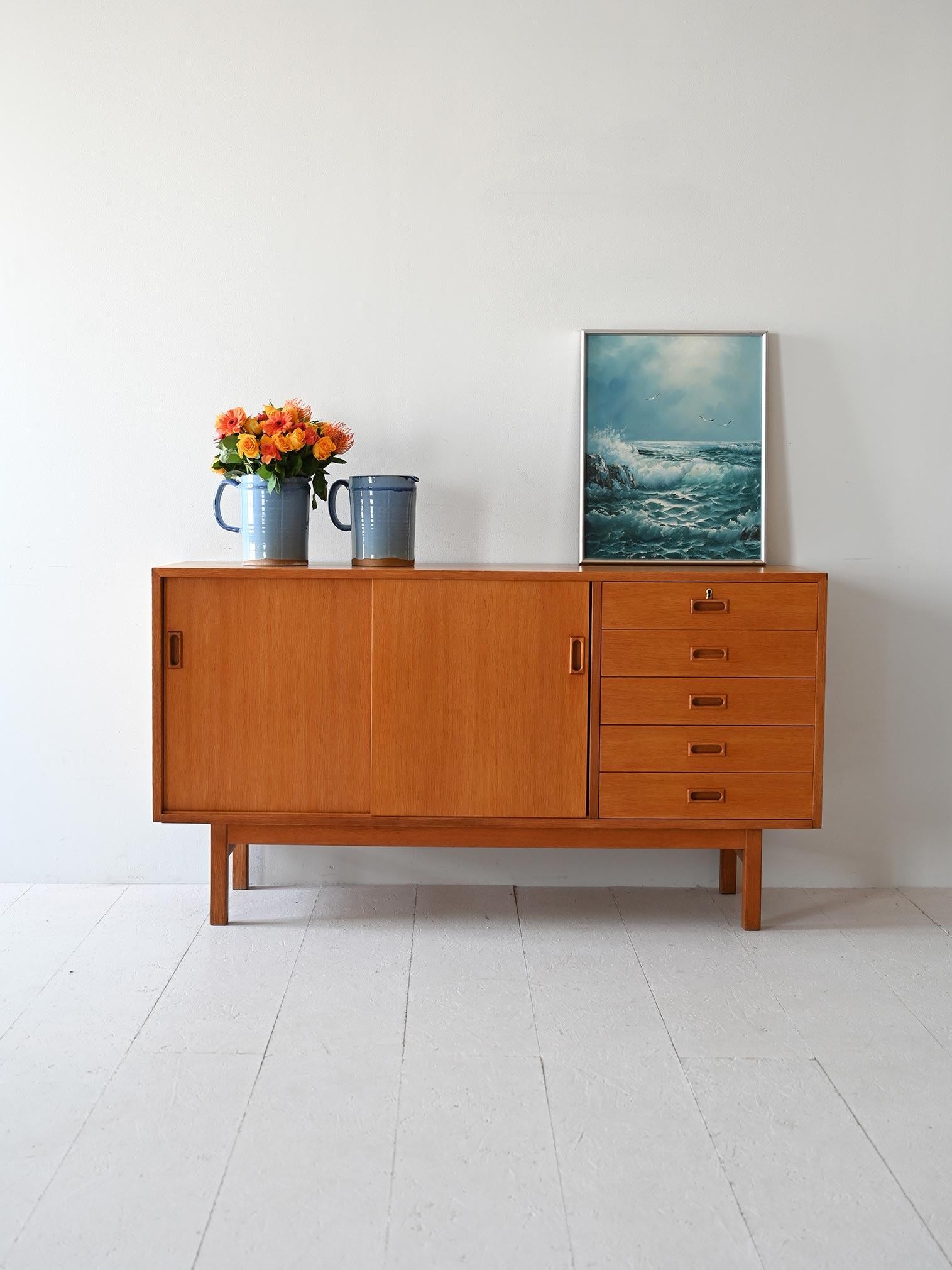 1960s teak sideboard with drawers and sliding doors. The handles are carved from wood. The first drawer is equipped with a lock. 

Inside the storage compartment is a shelf that divides the space. Also ideal as a TV cabinet. 

Good conditions.