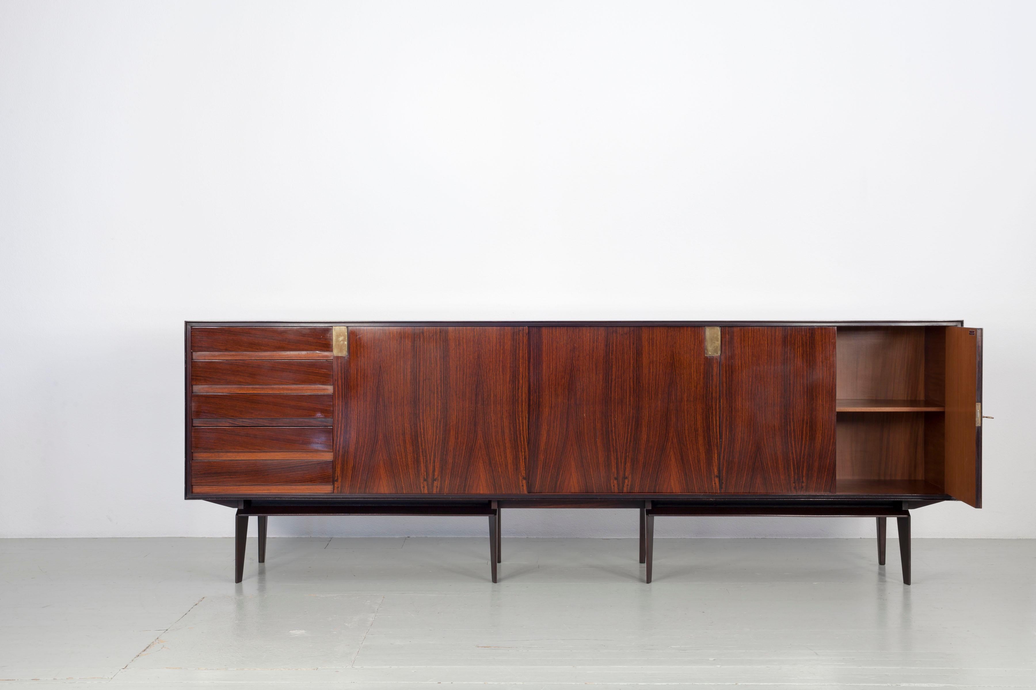 Elegant and timeless mid-century sideboard designed by Edmondo Palutari for Vittorio Dassi in the 1950s. It has solid and veneered rosewood structure with brass handles on sliding doors and carved wooden handles on drawers. The swing doors have an
