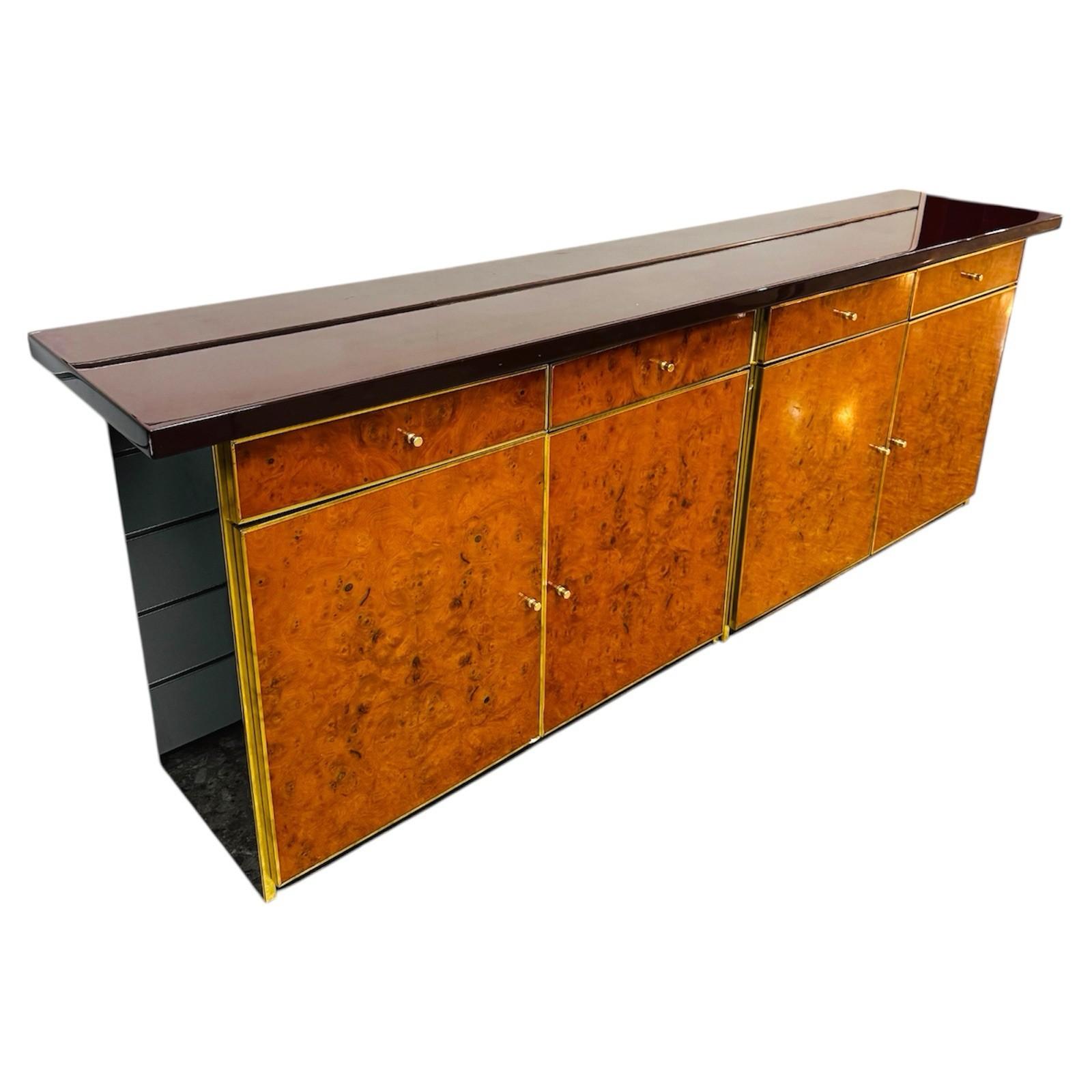 Elegante sideboard originale degli anni ‘70, emblema del modernariato italiano di lusso. La struttura presenta ante e cassetti in raffinata radica con venature calde e avvolgenti, incorniciati da profili in metallo dorato che donano un tocco di