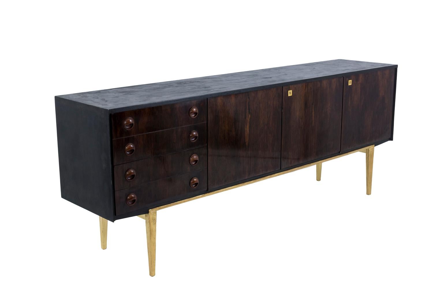 Sideboard in Rio rosewood veneered, opening by three front door leaves and four drawers standing on a gilt stand.
The three door leaves open on a large central compartment and a smaller lateral compartment, each one with a shelf. Drawers open thanks