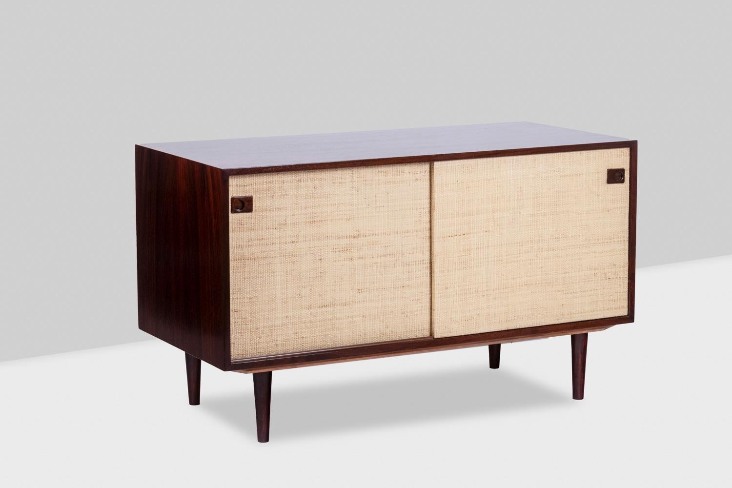 Rectangular rosewood sideboard opening with two sliding doors on the front, the doors covered in raffia. Spindle-shaped base. Rectangular shaped handles with a round shaped finish. Inside, a removable shelf in the center, the whole forming two