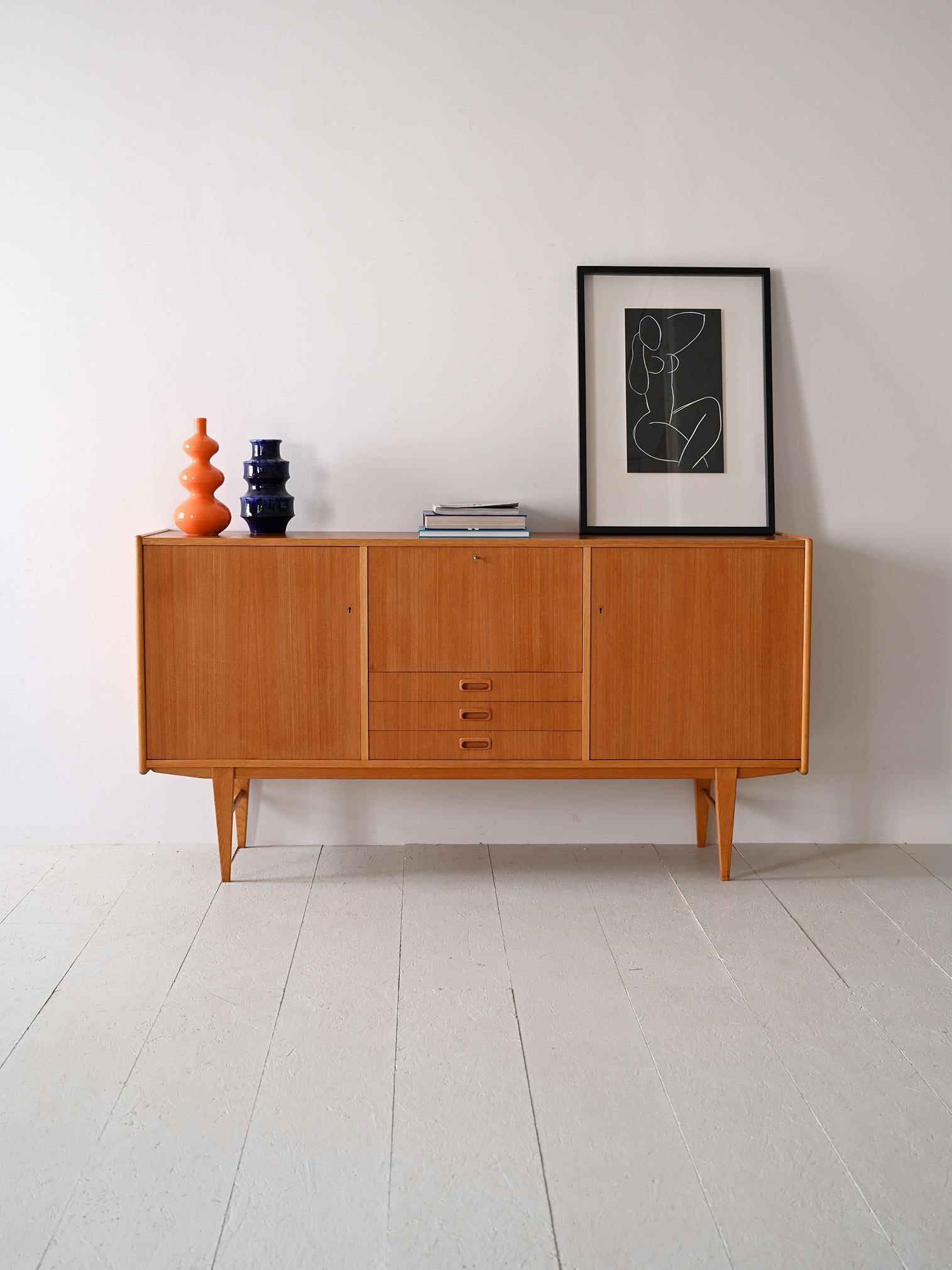 This 1960s Scandinavian oak sideboard is a testament to the functional design of the era. 
Equipped with two compartments with hinged doors and three central drawers, it offers a versatile storage solution. The flap door compartment adds a unique