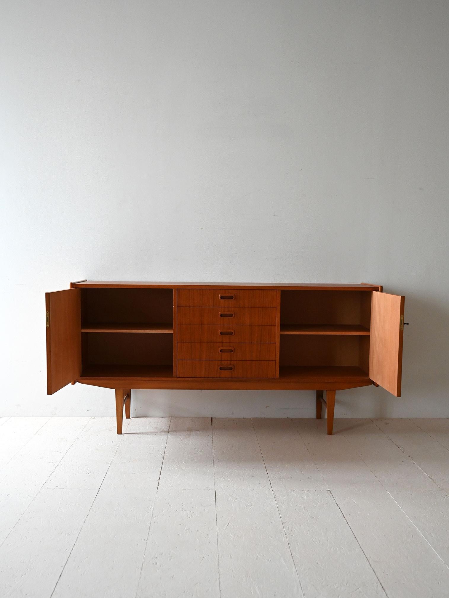 Sideboard in teak fine anni ’50 con due ante a battente, una mensola interna e sei cassetti centrali con maniglie intagliate.

Linee sobrie e struttura solida per questa sideboard scandinava in teak risalente alla fine degli anni ’50. Le due ante
