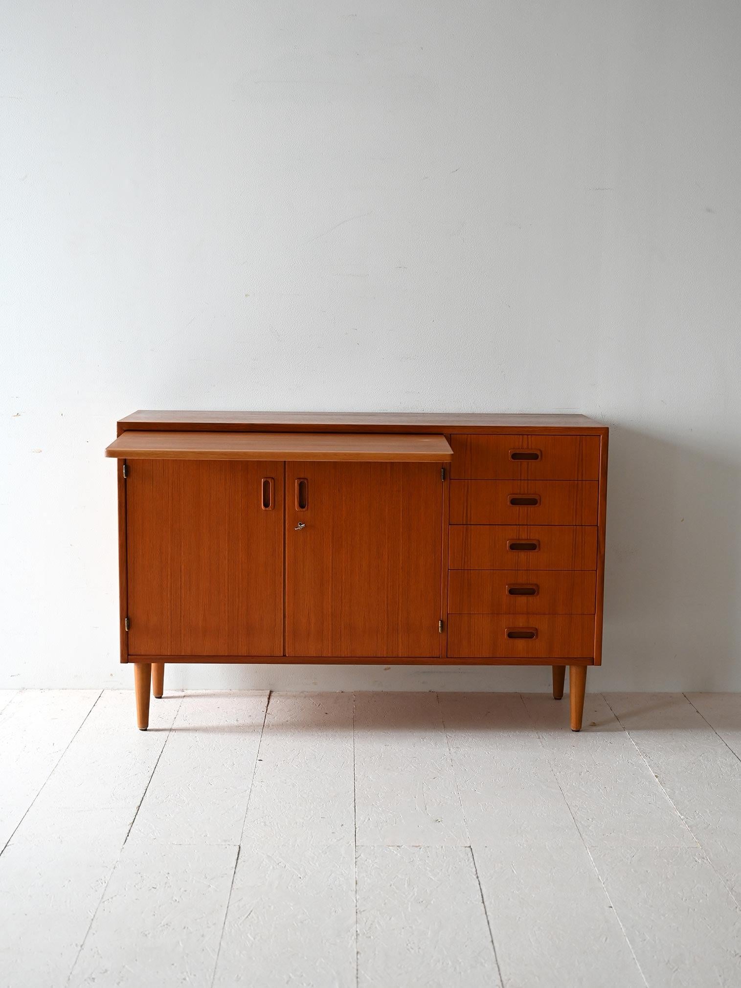 Late 1950s teak sideboard with five drawers, doors with carved handles, and concealed writing desk.

Compact and balanced structure made of teak with uniform grain. The cabinet features five drawers with carved handles and a compartment closed by