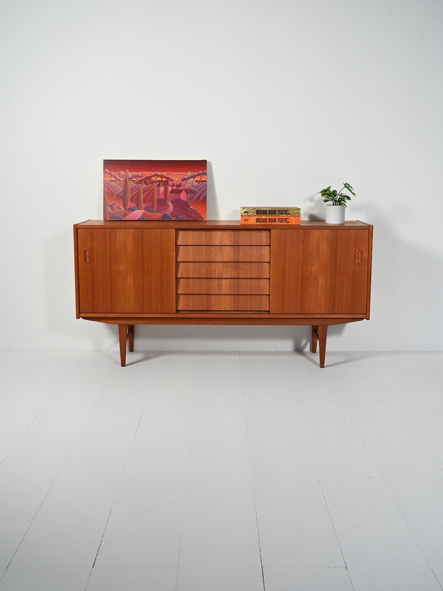 Credenza in teak con ante scorrevoli e blocco di cassetti centrali a listelli. Gambe rastremate, venatura leggera e tono caldo.

Linee affilate e proporzioni leggere esaltano il teak caldo di questa credenza anni 50/60. Le ante scorrevoli con