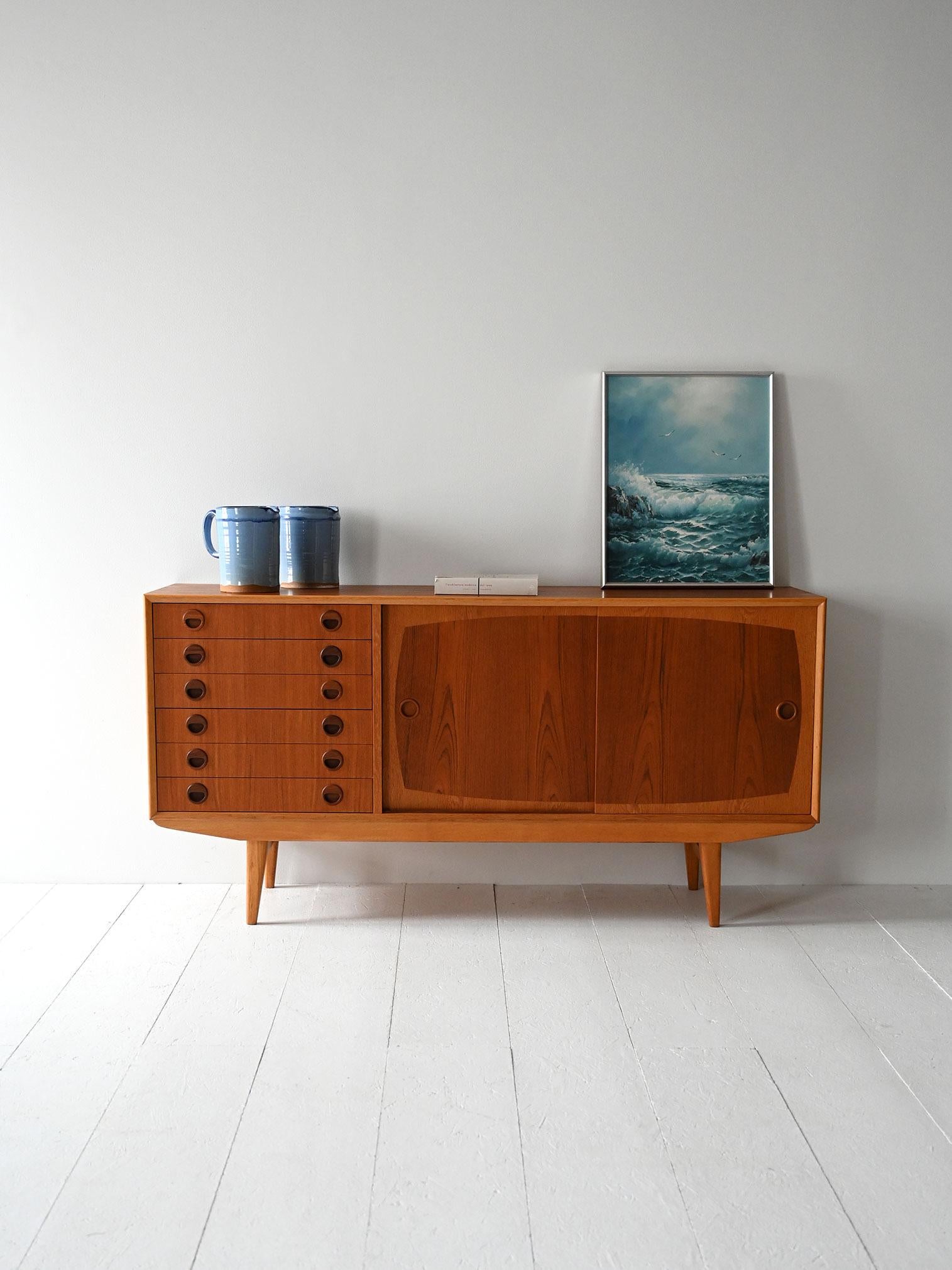 Sideboard in teak e rovere con sei cassetti e due ante scorrevoli. Maniglie rotonde intagliate e doppia essenza.

Realizzato tra la fine degli anni ’50 e l’inizio dei ’60, questo sideboard scandinavo gioca sull’alternanza tra teak e rovere. Sei