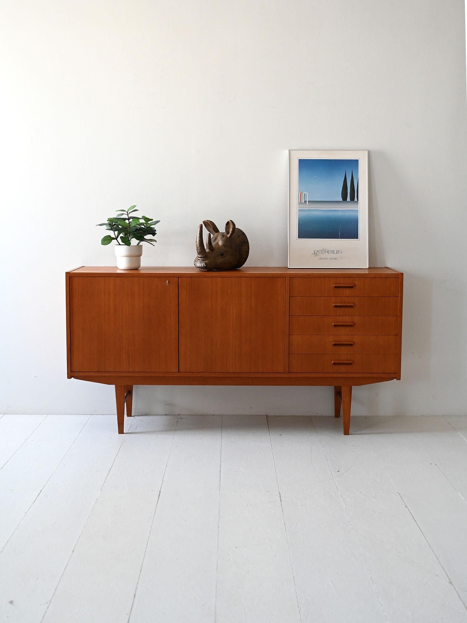 Buffet vintage en teck avec porte à double charnière, compartiment de rangement interne et cinq tiroirs latéraux avec poignées elliptiques encastrées.

Buffet scandinave des années 1960 en teck, caractérisé par des lignes épurées et un équilibre