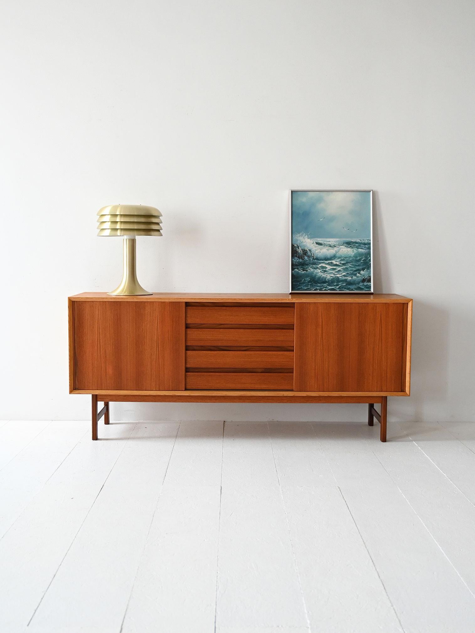 Sideboard anni ’60 in teak con ante scorrevoli, vano interno con mensola e cassettiera centrale a latta.

Sideboard scandinavo in teak databile alla fine degli anni ’60. Le ante scorrevoli laterali nascondono vani con mensole interne, mentre al