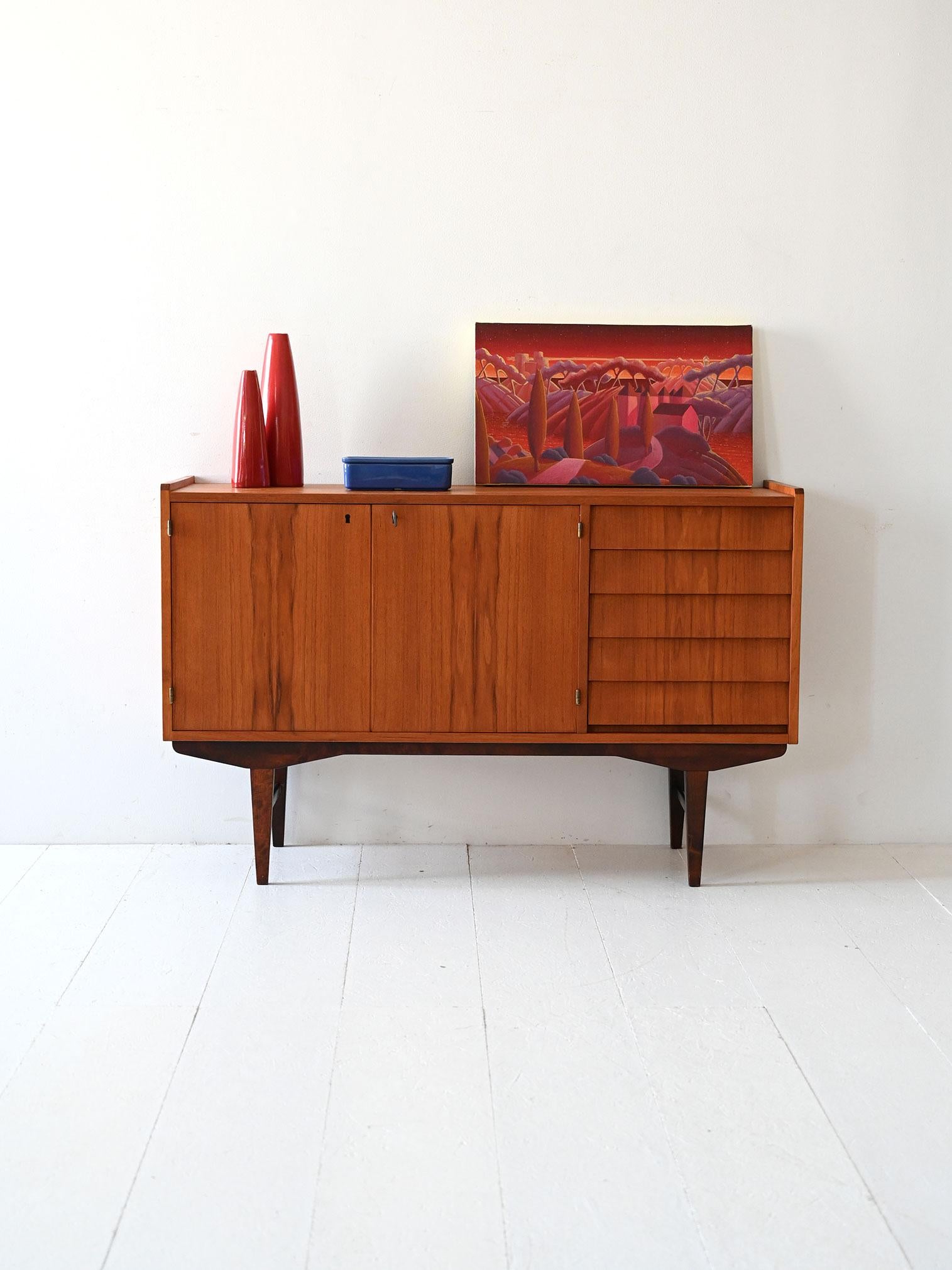 Sideboard in teak con doppie ante a battente, vano con mensola interna e cassetti a profilo a tegola. Gambe rastremate in teak scuro.

Sideboard scandinavo vintage in teak realizzato negli anni ’60, caratterizzato da doppie ante a battente con vano