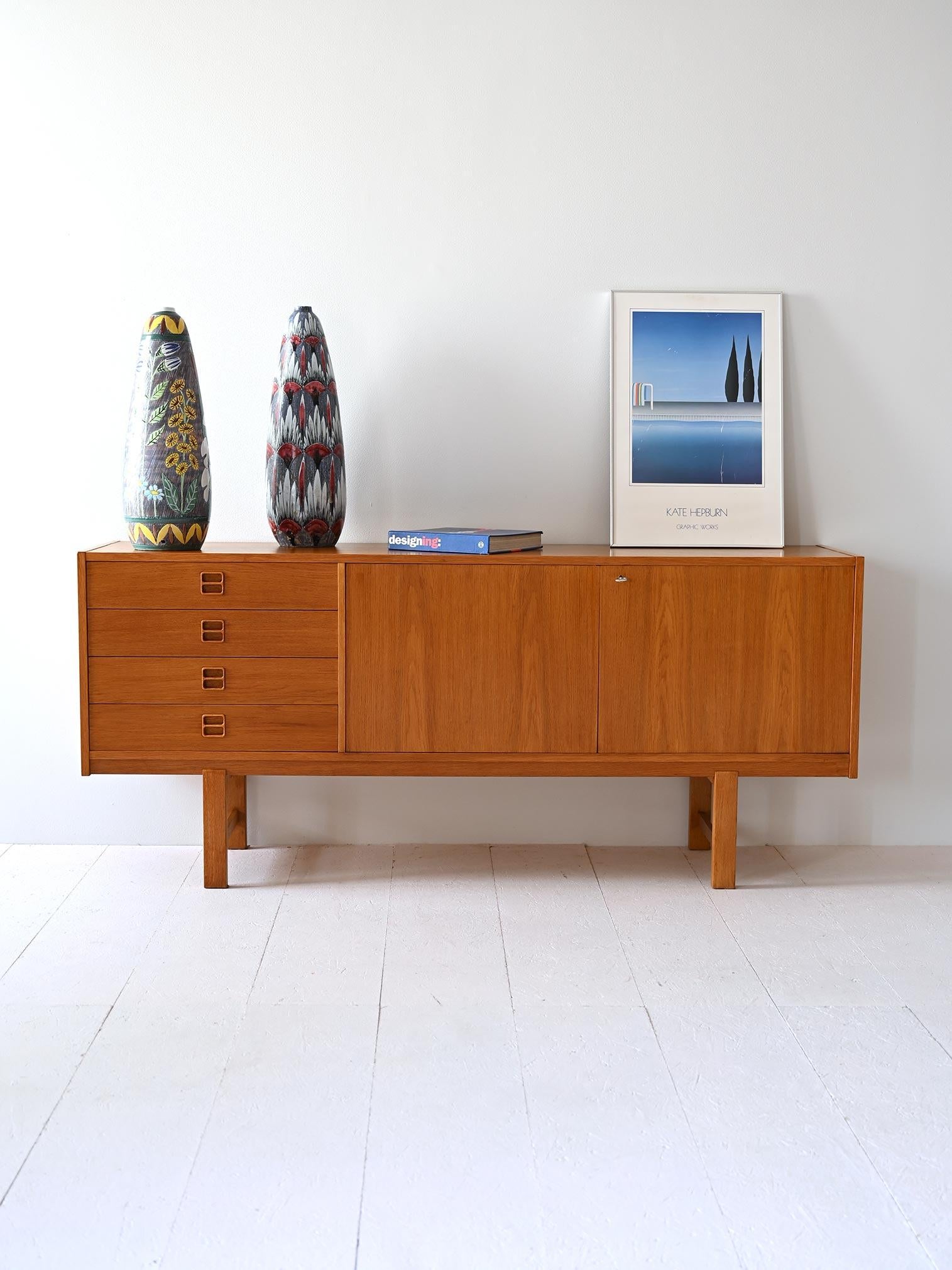 Scandinavian sideboard with minimalist design. On one side are four drawers with wood-carved handles, and on the other side is a large compartment is closed by two lockable hinged doors. 

Square legs lift the sideboard off the ground, giving the
