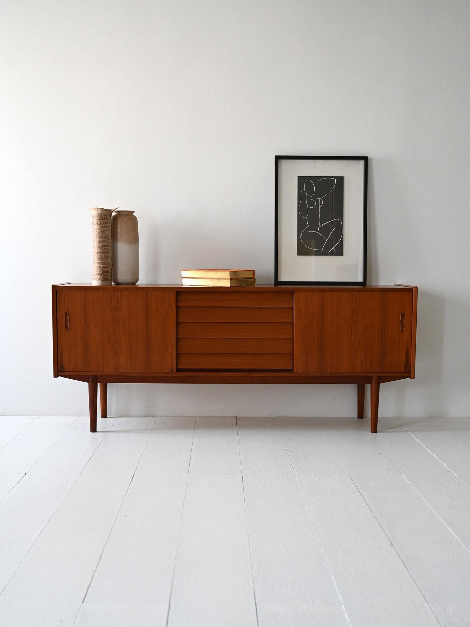 Teak sideboard with sliding doors and center tile drawers. Scandinavian design by Nils Jonsson.

Teak sideboard designed by Nils Jonsson for Hugo Troeds in the 1950s. The structure consists of sliding side doors with an interior shelf and central