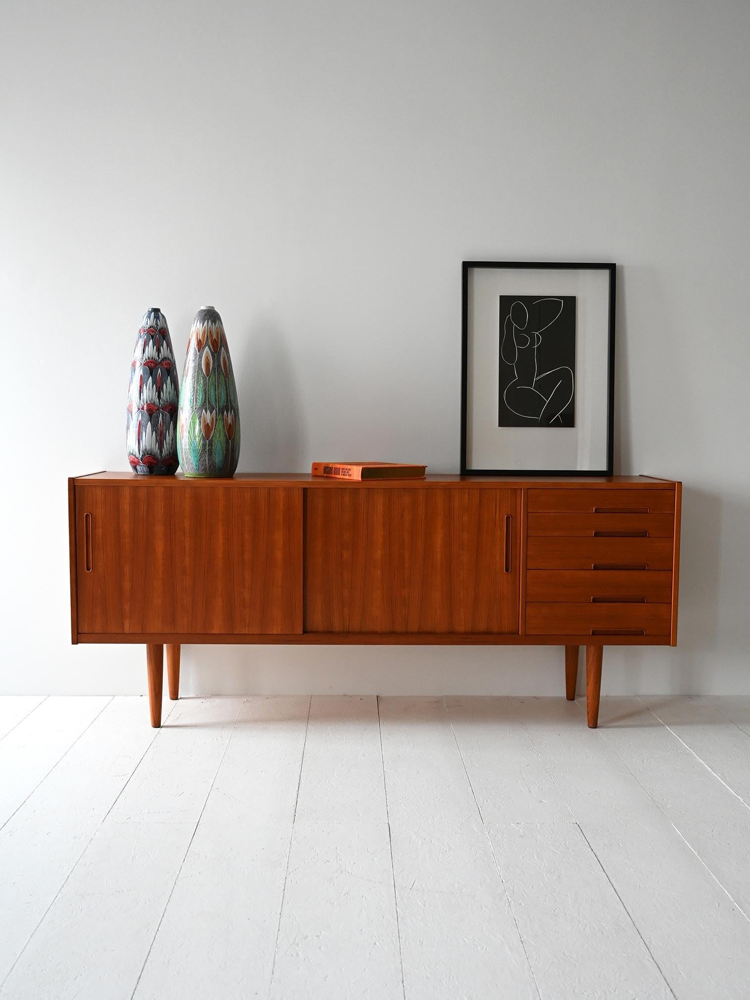 Vintage 1960s teak sideboard with sliding doors, five drawers and carved handles.

Elegant Scandinavian sideboard designed by Nils Jonsson for Bra Bohag, made in the early 1960s. Made of teak, with five aligned drawers and two sliding doors