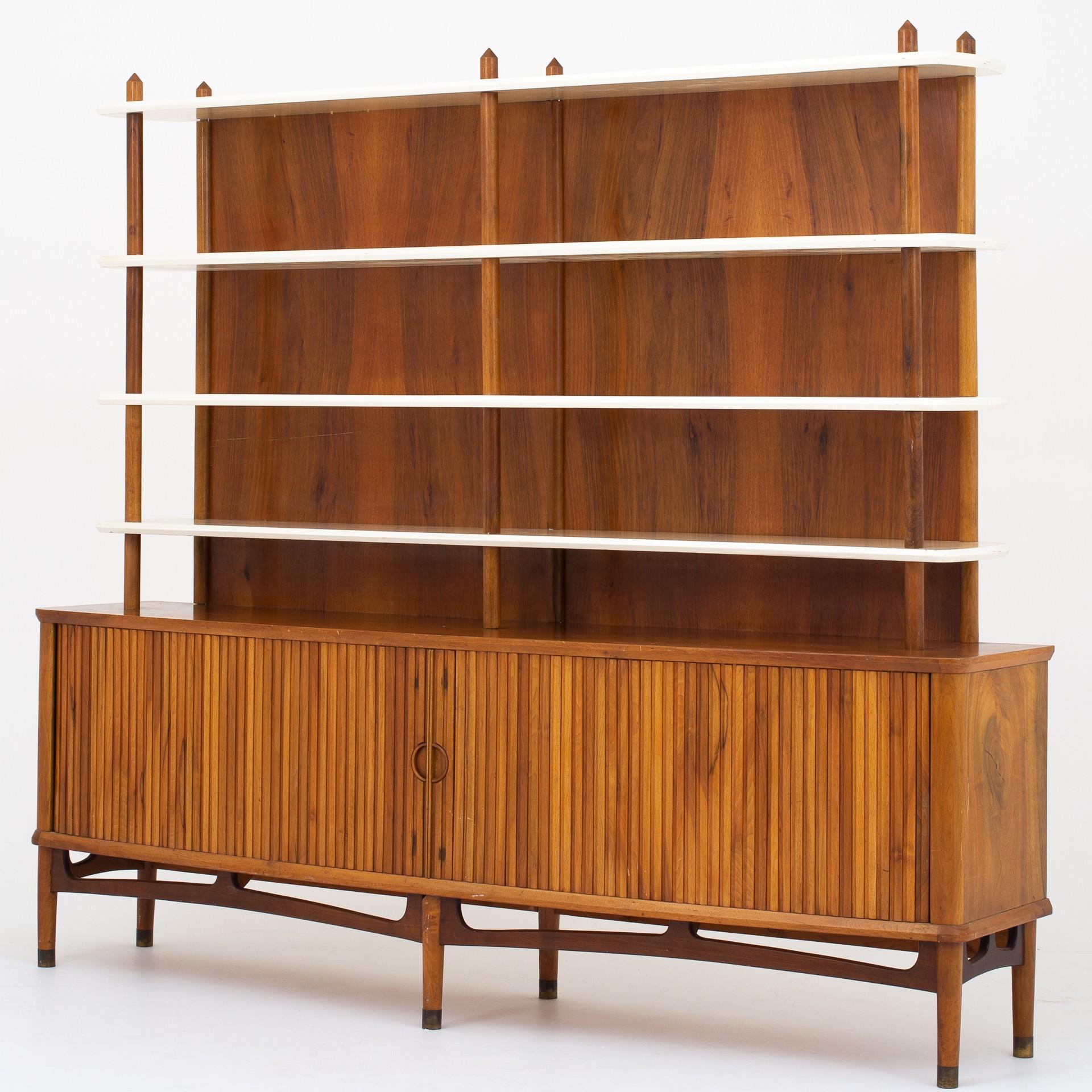 Sideboard with shelving, varnished walnut veneer, tambour doors. Top section with white-lacquered shelves and tapered pillars, base with slightly bowed front and two tambour doors enclosing shelves and sliding trays. Maker A. Andersen & Bohm.
