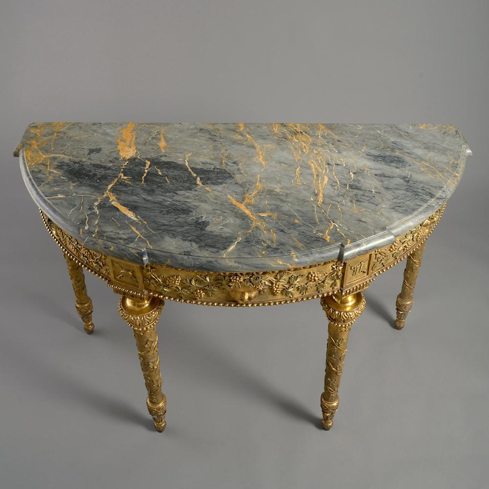 A fine Tuscan (Siena) carved giltwood demilune side table, circa 1790.

With original eared moulded grey Siena (Breddolato Senese) marble top. The frieze carved with trailing vines centred by a Bacchic mask on turned and tapering legs entwined by
