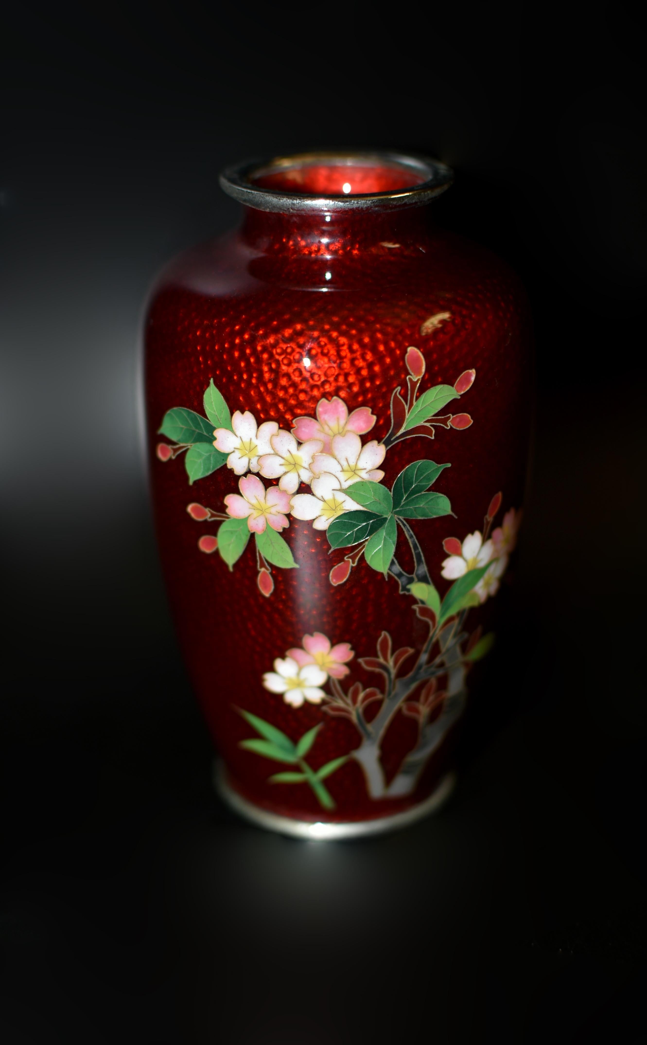 Un raro vaso di cloisonne Ando Jubei ginbari. Sul corpo due rami di fiori di ciliegio bianchi e rosa in piena fioritura e in forma di bocciolo con fogliame verdeggiante, lavorati minuziosamente, su uno splendido sfondo rosso metallizzato. Il Ginbari