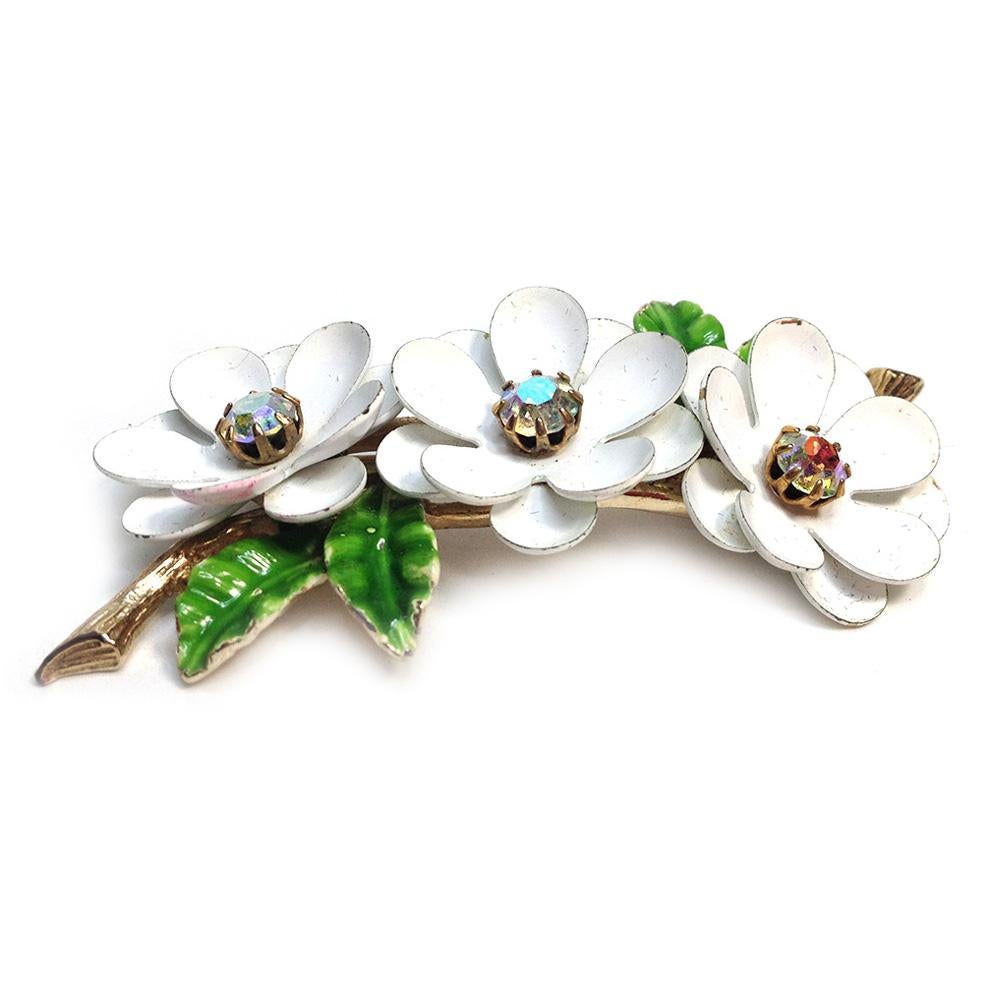 This signed Art white enameled flower brooch is from the late 1950s. There is a row of three white enameled flowers with AB rhinestones in centers and riveted on a gold plated twig with green enameled leaves. This triple dogwood brooch will take you