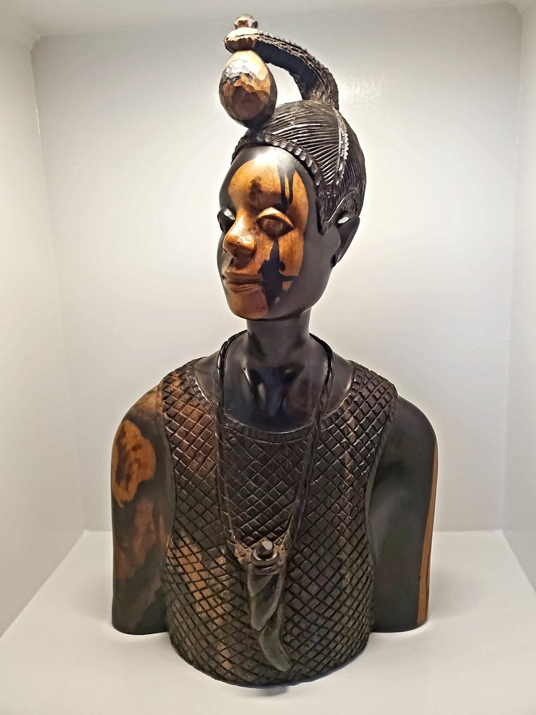 This beautifully carved bust by Felix Ogbe Ozo is of a Nigerian man wearing a textured sleeveless top with a horn-shaped flask around his neck. His hair is dressed with a large ornament in the traditional suku style of the Yoruba Peoples. The wood