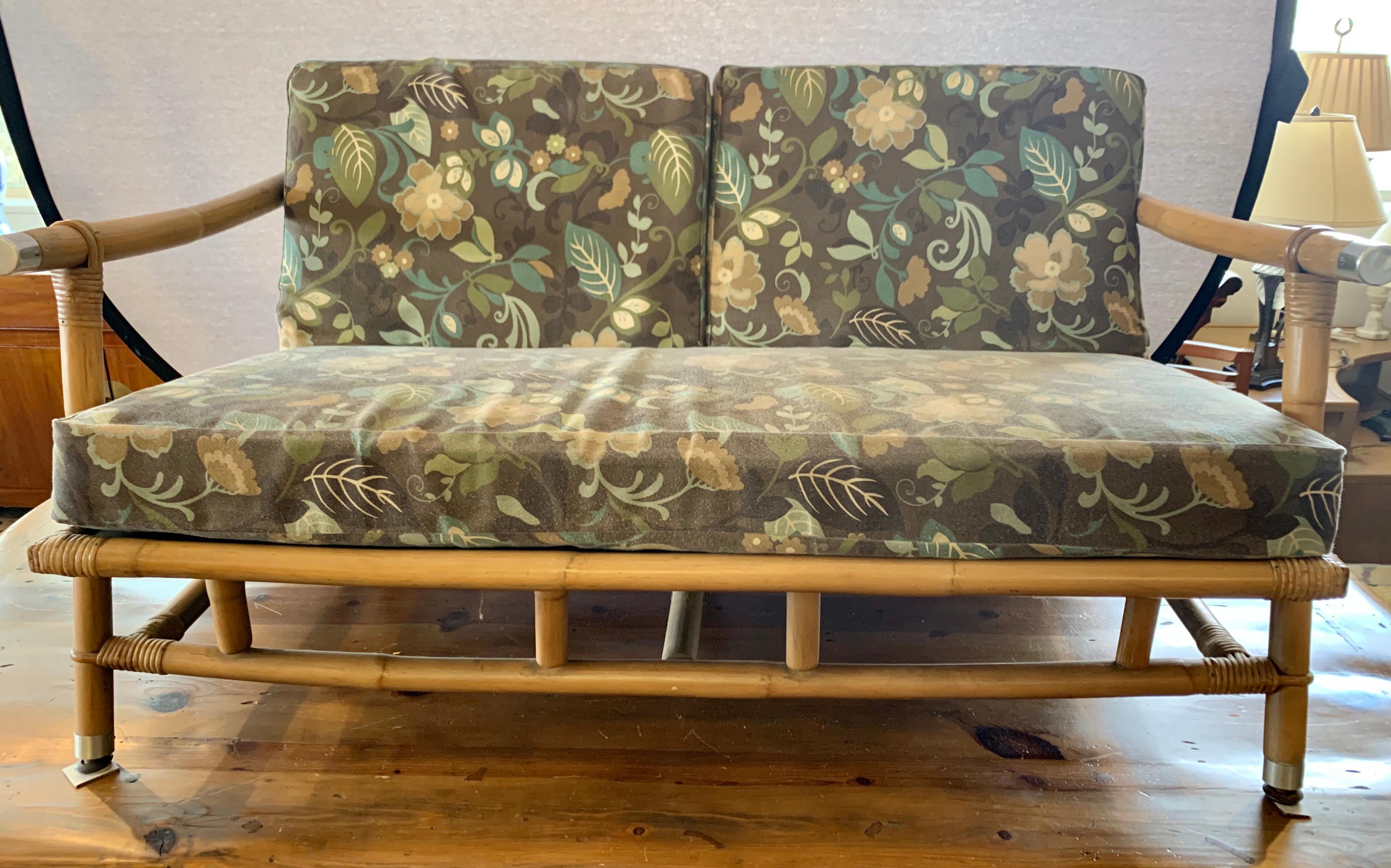 A coveted vintage Campaign style loveseat made by Ficks Reed and designed by John Wisner, circa 1954. Stout rattan and bamboo frame with a beautiful raffia seat back. Handsome solid metal accents mark the arms. Fabulous design, quality and