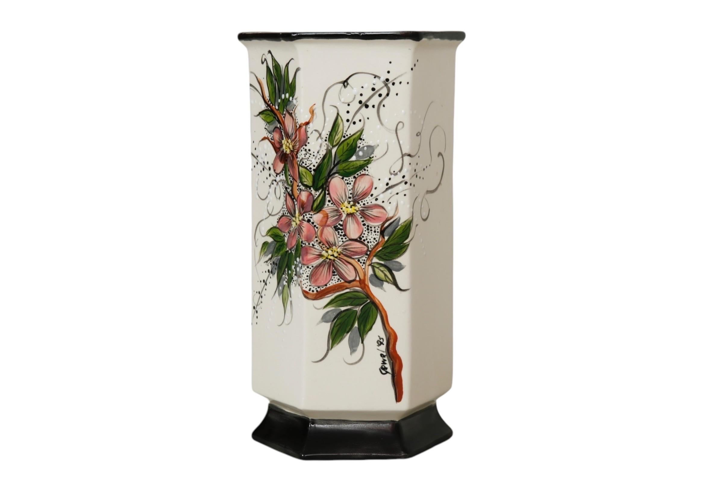 Un vaso decorativo esagonale in ceramica, dipinto a mano con un delicato spruzzo floreale di fiori rosa nella parte anteriore, impreziosito da punti bianchi e neri in rilievo su sfondo bianco. Il labbro e la base sono neri e il vaso è firmato e
