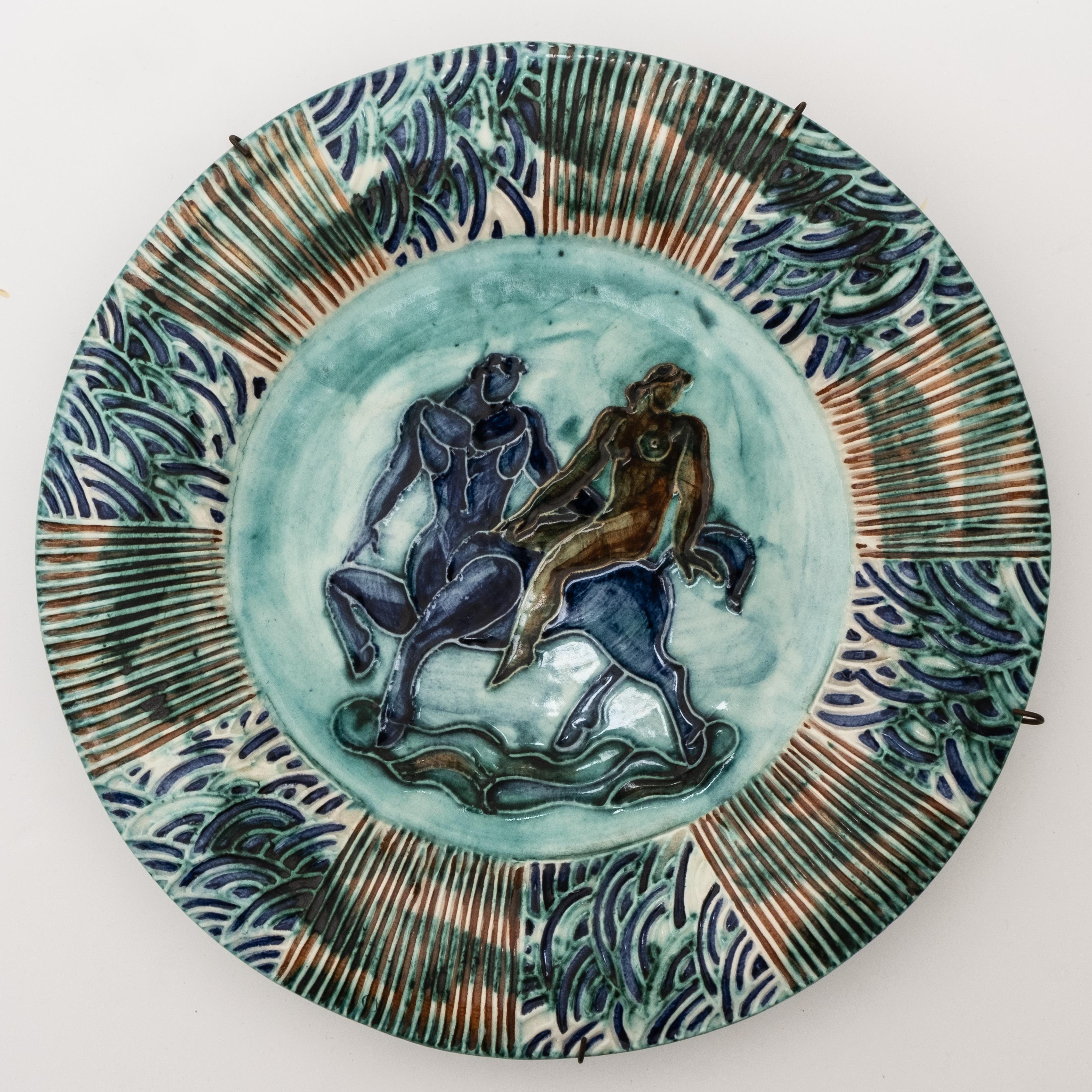 Assiette en céramique émaillée Art Déco, présentant une scène figurative centrale avec un centaure et une femme nue, entourée d'une bordure émaillée texturée et polychrome dans des tons de bleu et de vert, signée 