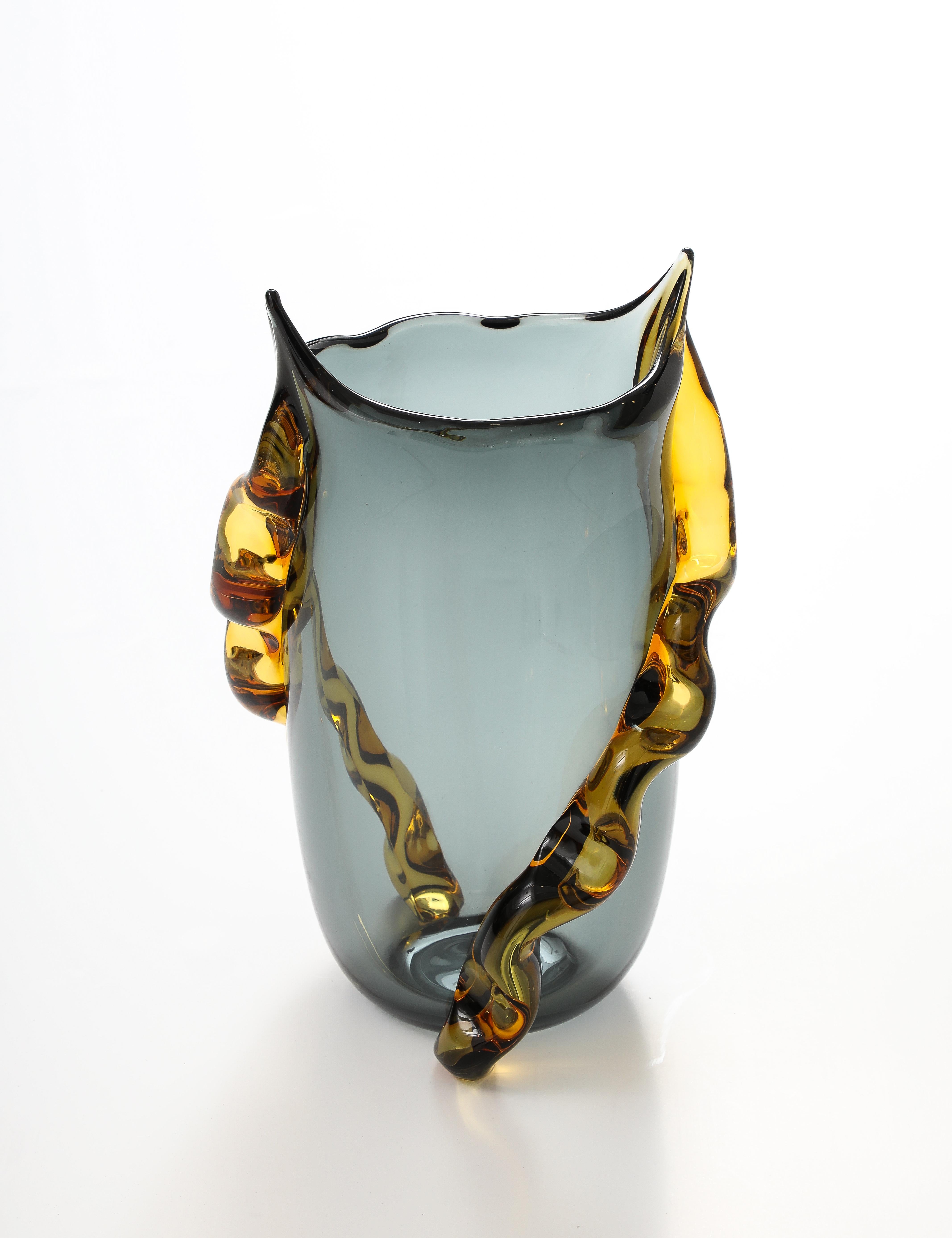 Un jarrón grande y sustancioso, soplado a mano y fabricado con maestría en Murano (Italia), con un llamativo cuerpo translúcido de color gris marengo y una silueta asimétrica que realza su presencia escultural. Dos gruesas asas de ámbar de forma