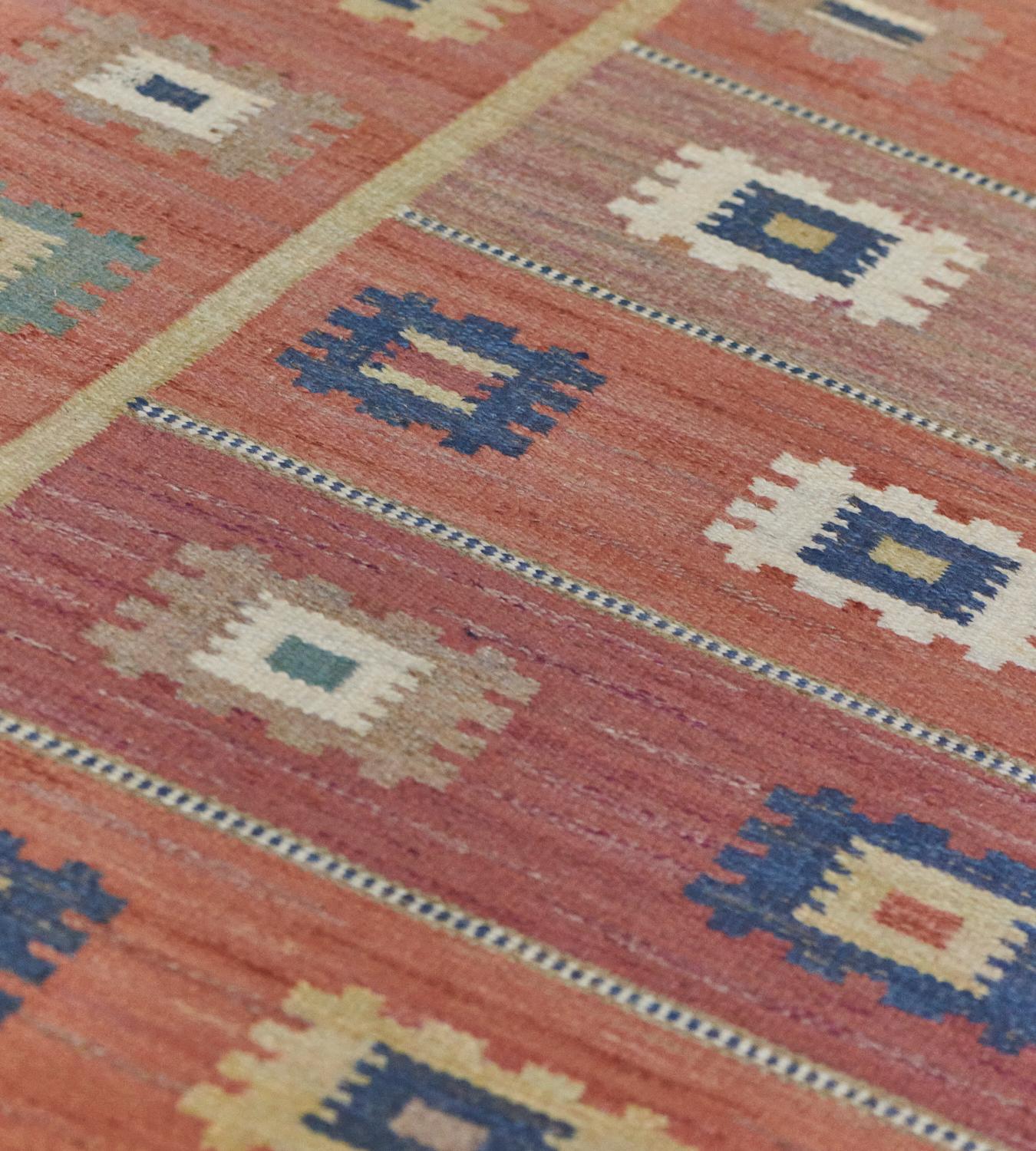 Ce tapis suédois vintage tissé à la main présente un champ orange sang avec des bandes complémentaires alternées d'icônes carrées dentelées polychromes, dans une bordure orange similaire. Signé par l'atelier d'origine.

Sur le maître tisserand