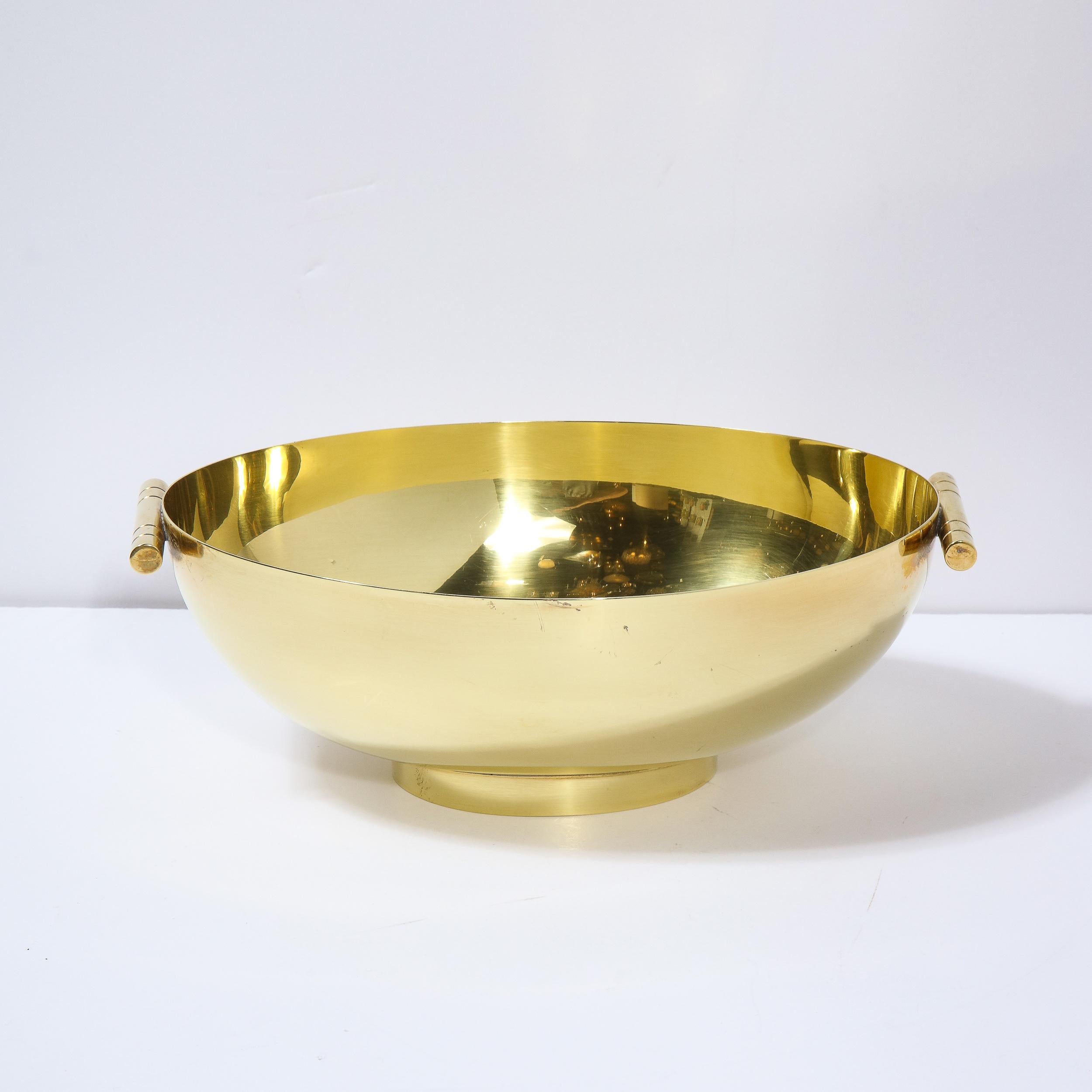 This elegant Mid-Century Modern polished brass bowl was designed by Tommi Parzinger for Dorlyn Silversmiths in the United States circa 1960. It features a concave oval body with a cylindrical banded handle on each side in lustrous brass. With its