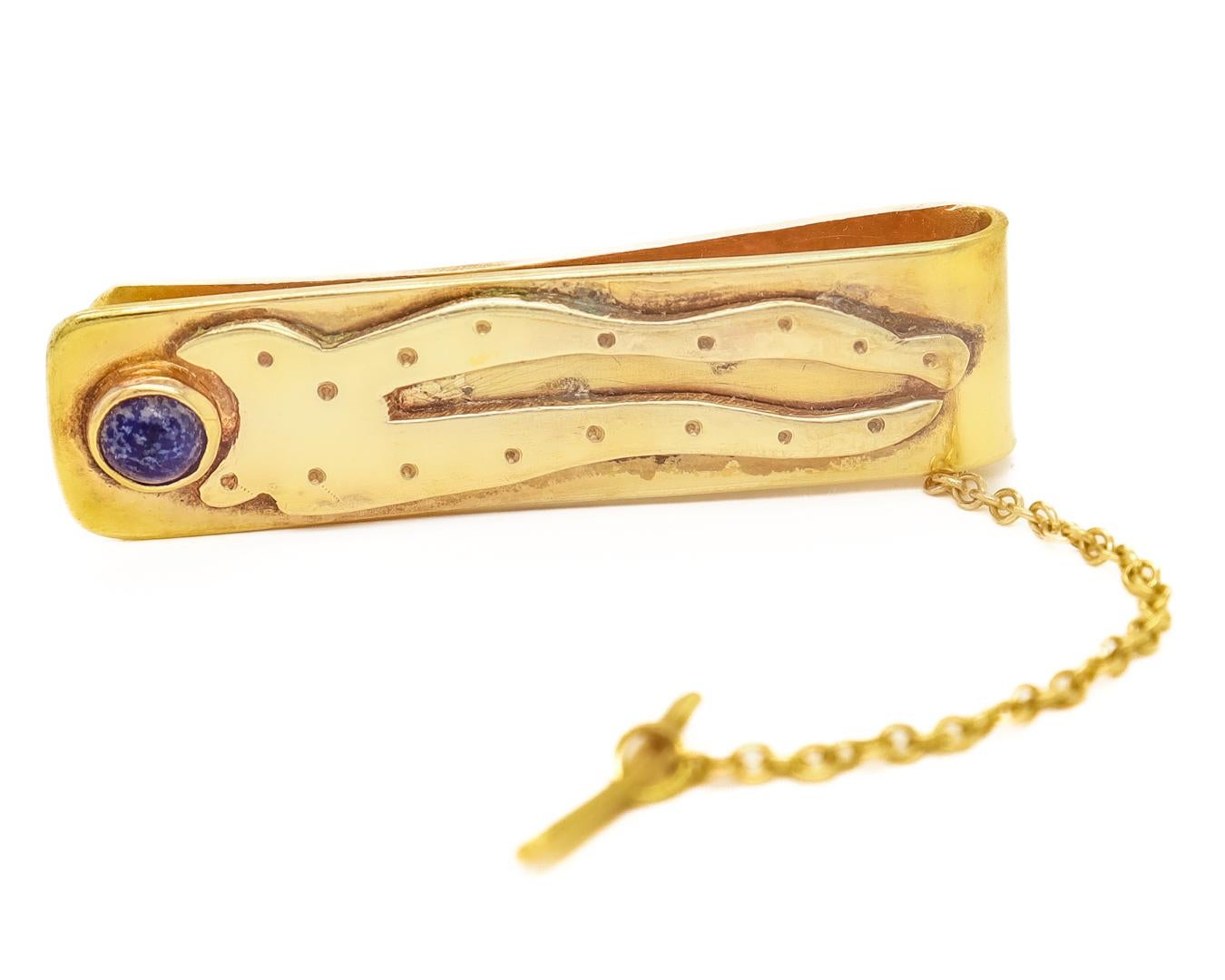 A fine signed Modernist tie bar.

By Sam Kramer.

In 14k gold.

Bezel-set with a round lapis lazuli cabochon and with an applied decoration to the head.

Set with a safety chain and a button bar. 

(The piece could easily also double as a money