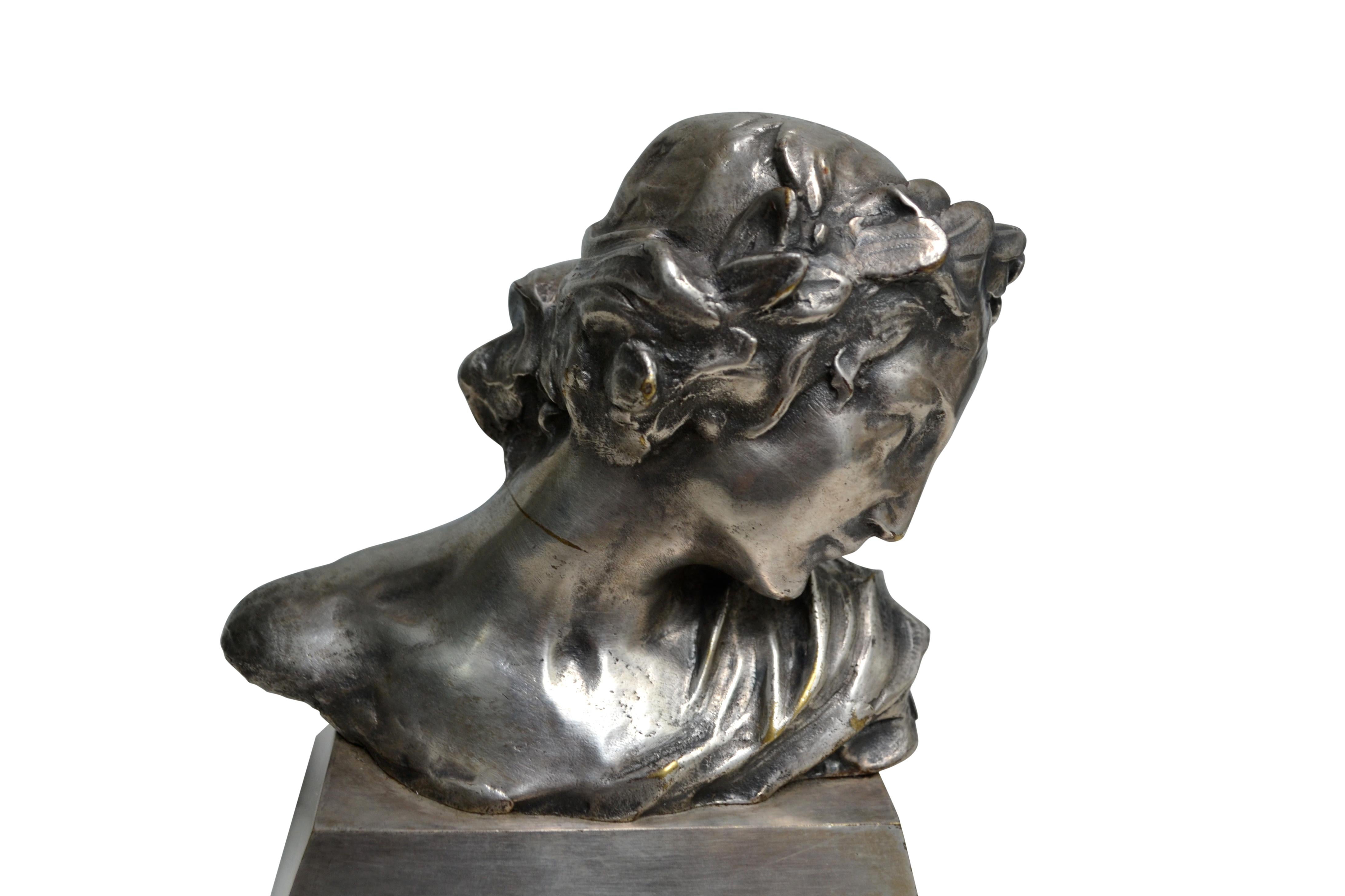 A beautifully cast Art Nouveau silvered bronze bust of a classically draped and coiffed woman wearing a laurel leaf crown reminiscent of a Roman Empress. The figure is shown looking down demurely over her left shoulder in what is a very neoclassical